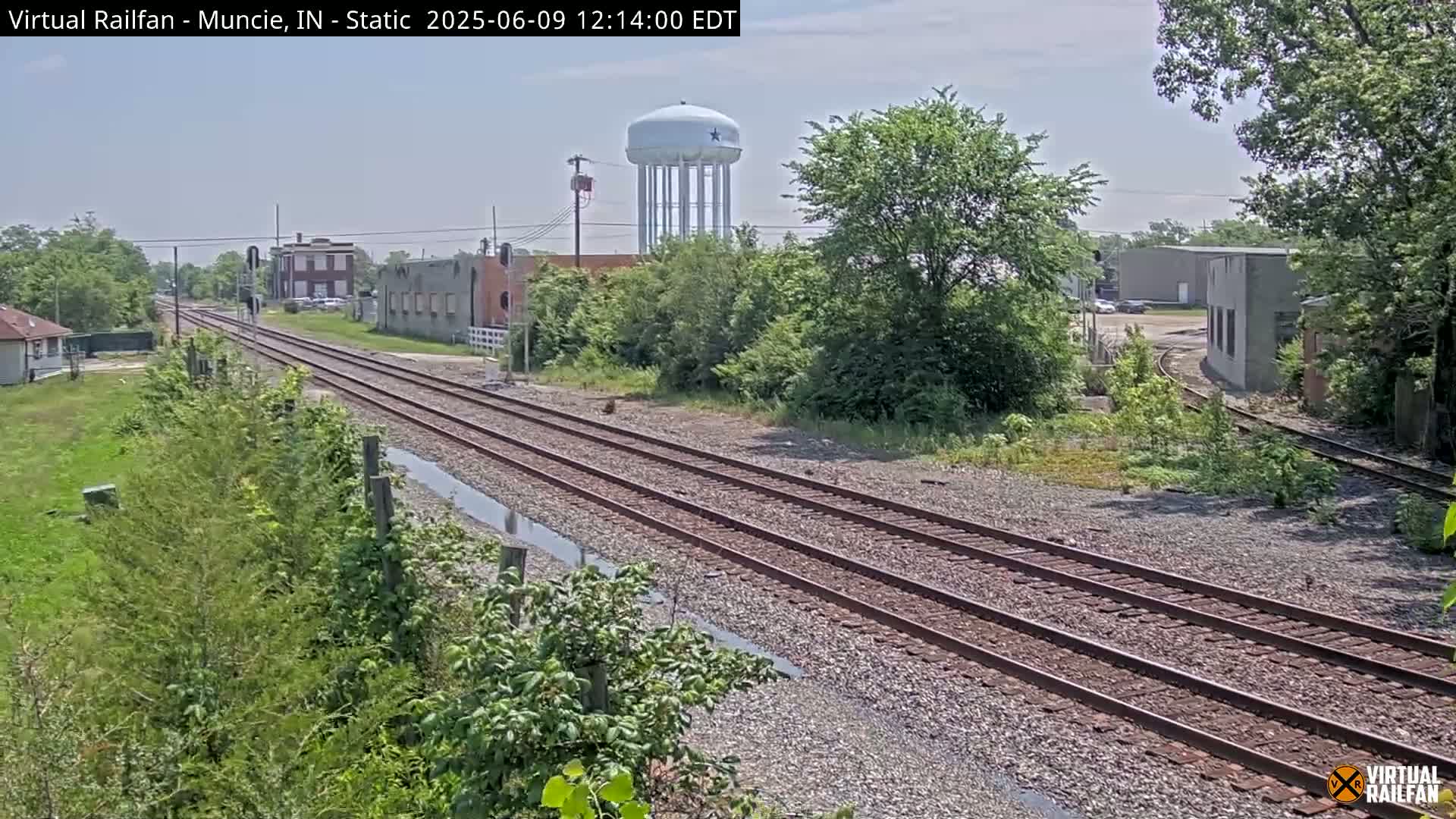 A view of railroad tracks on a partly sunny day, with buildings, greenery, and a water tower in the background.