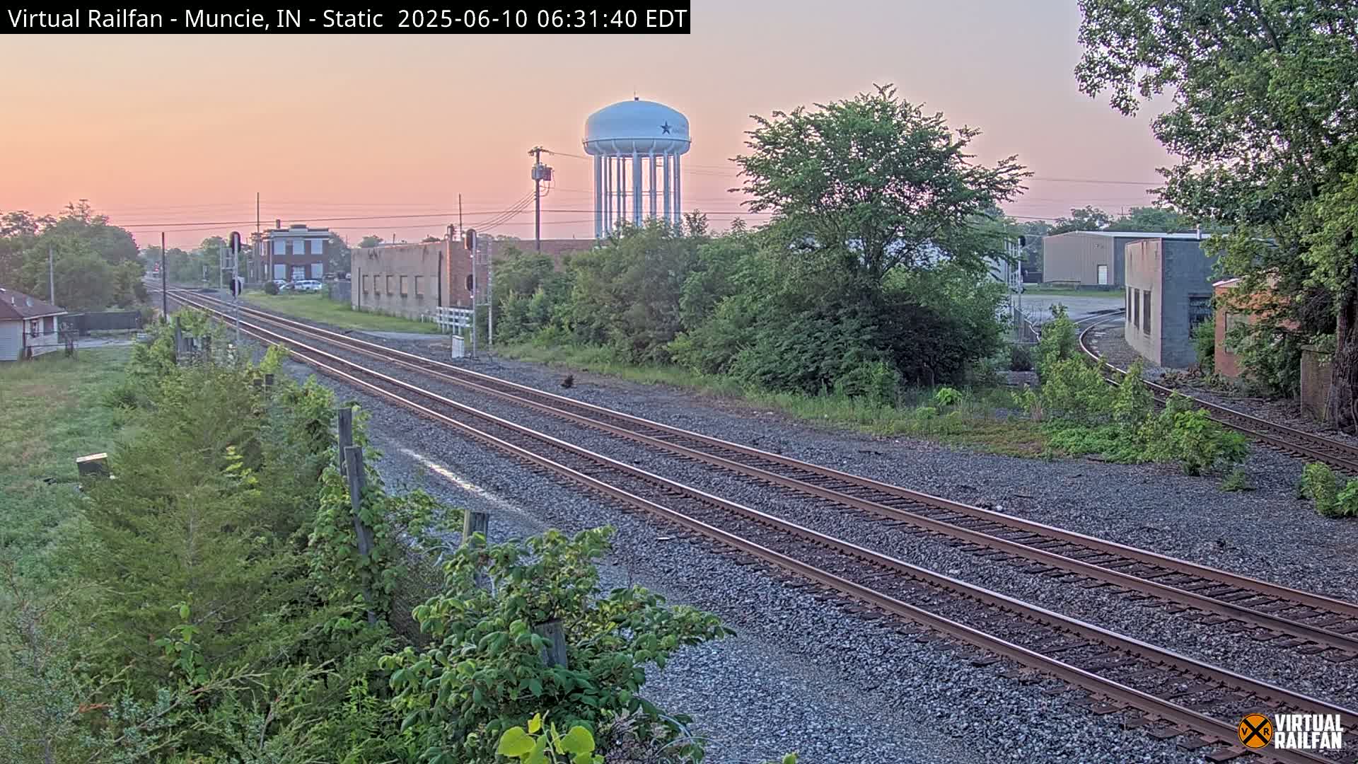 Triple railroad tracks extend into the distance under a pinkish dawn sky, past industrial buildings and a water tower, amidst green vegetation.