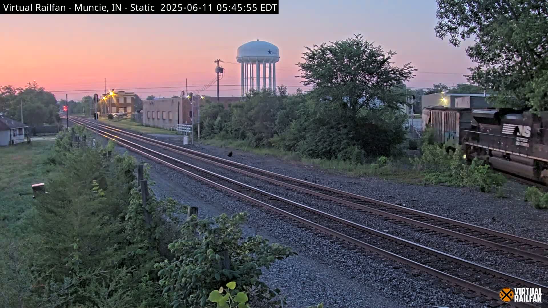 A sunrise illuminates a railroad track passing through a town with a water tower visible in the distance, a freight train partially visible on the right, and vegetation lining the sides of the tracks.