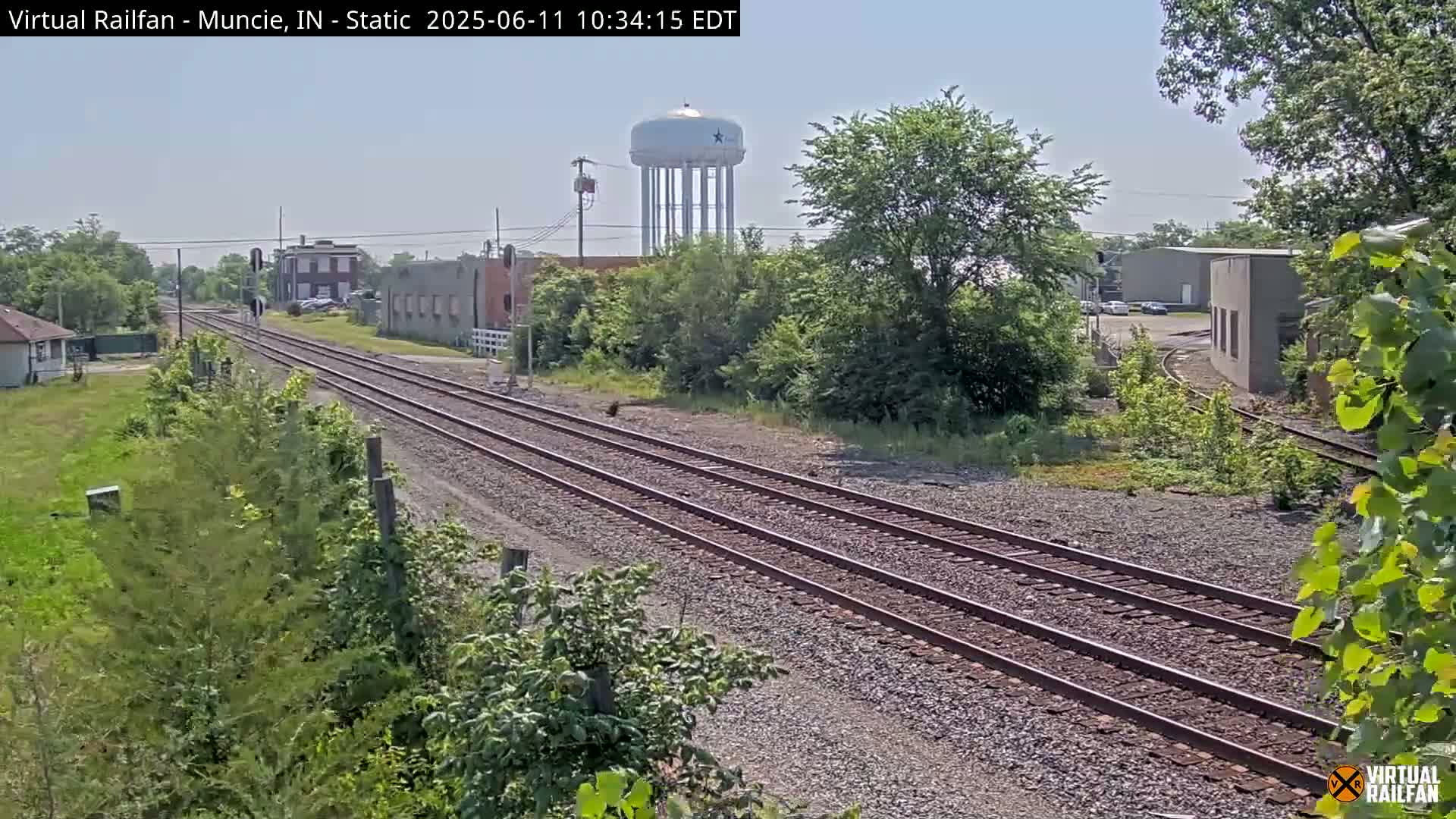 A view of train tracks on a sunny day, with some buildings and a water tower visible in the distance.