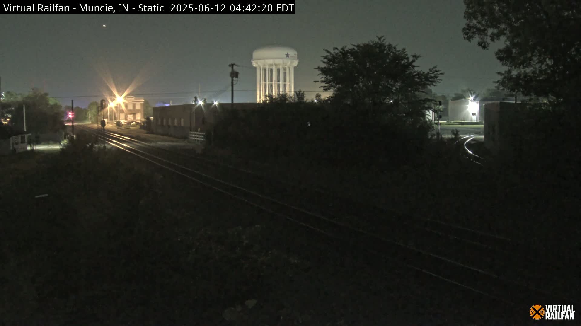 A nighttime view of railroad tracks leading toward a water tower and several buildings under a dark, possibly foggy sky.