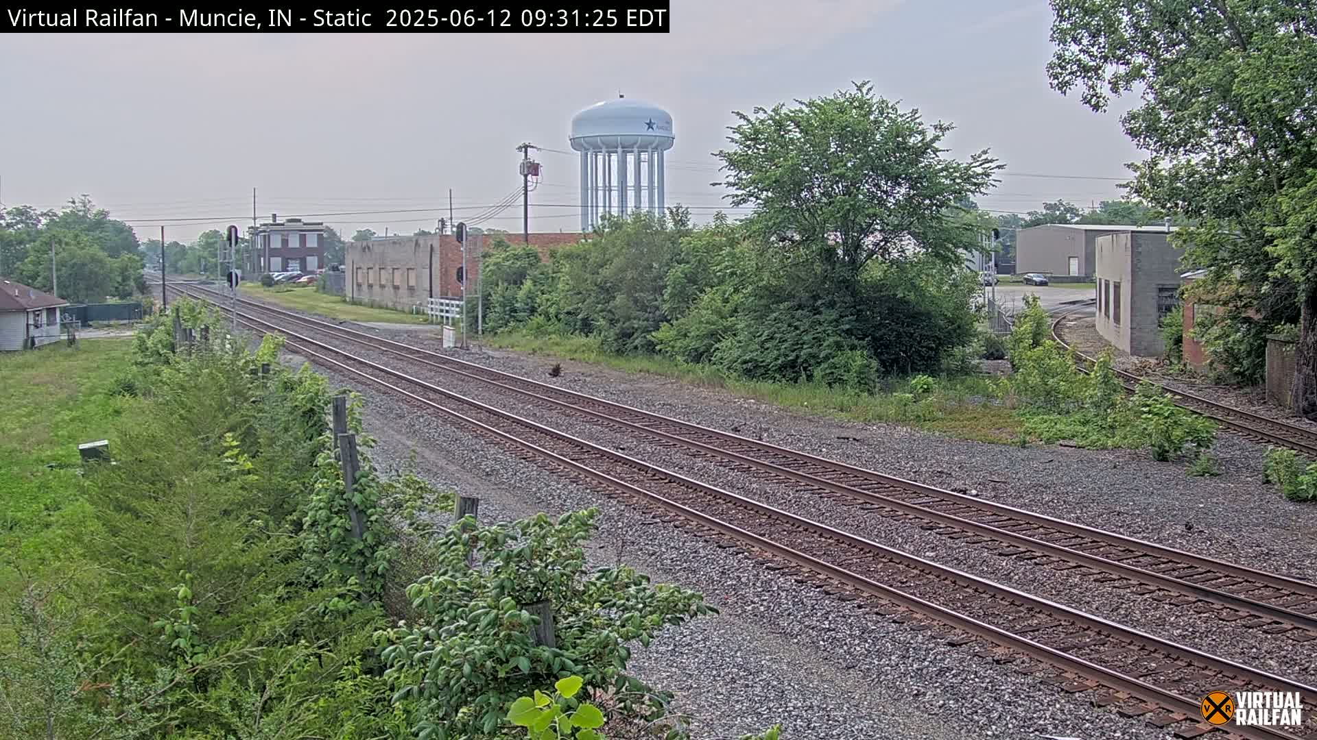 A view of railroad tracks on an overcast day, with a water tower visible in the distance, alongside some industrial buildings and lush green vegetation.