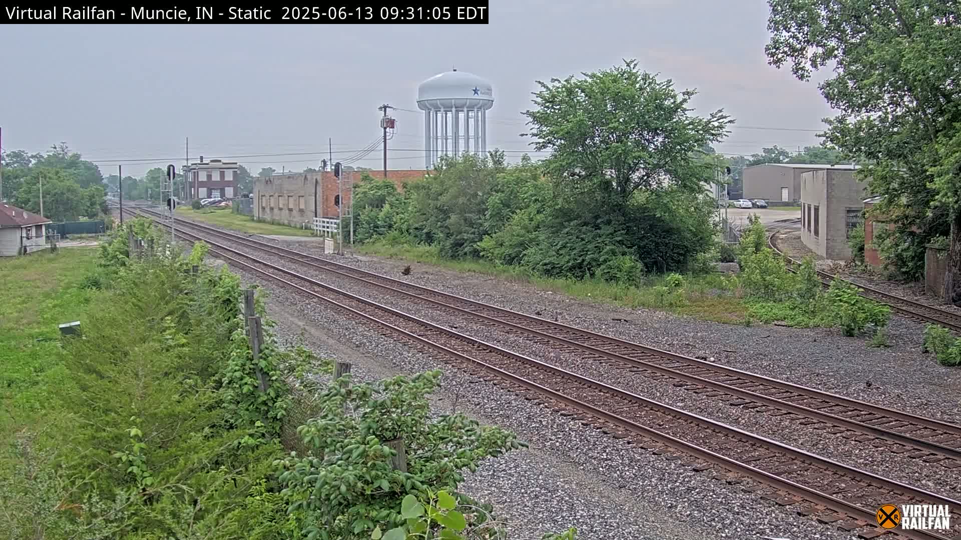 A view of a railway track running through an area with buildings, trees, and a water tower under an overcast sky.