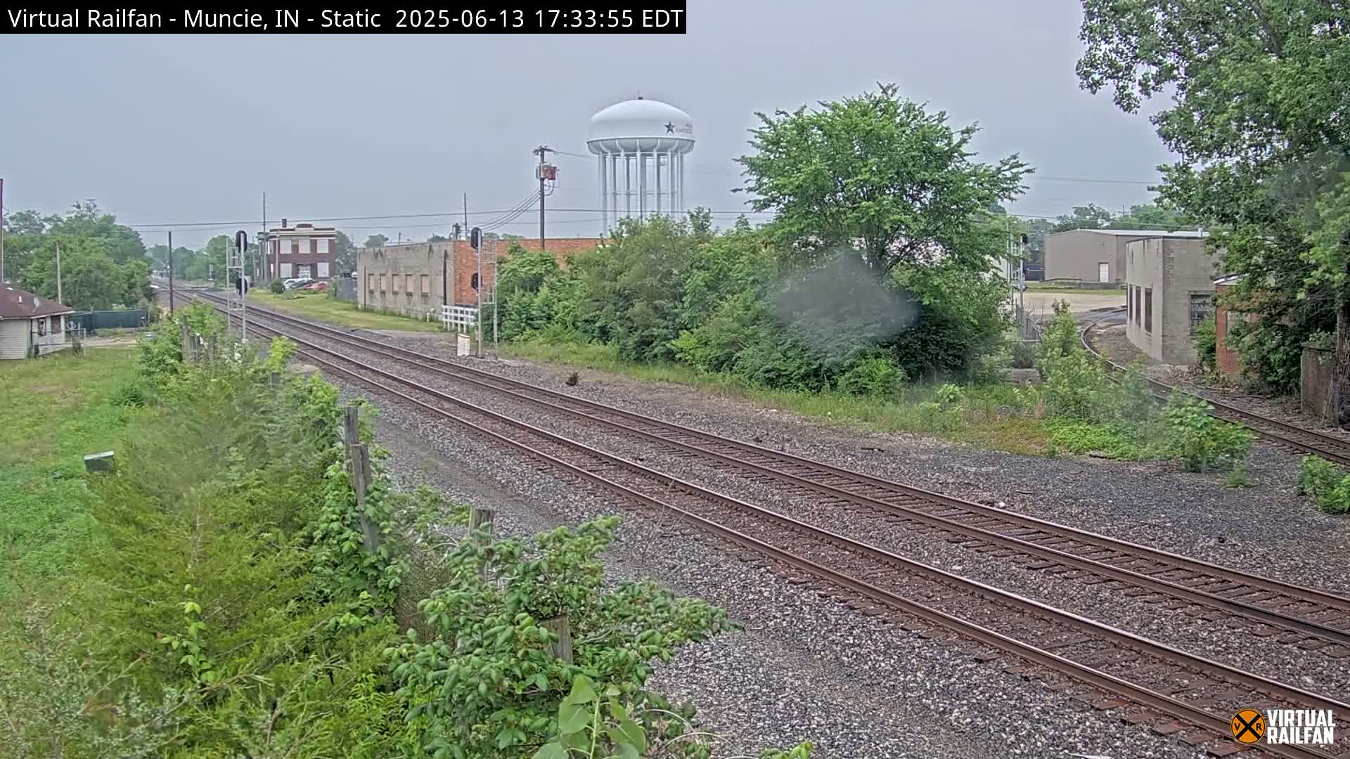A view of railroad tracks on an overcast day, with a water tower and industrial buildings visible in the background.