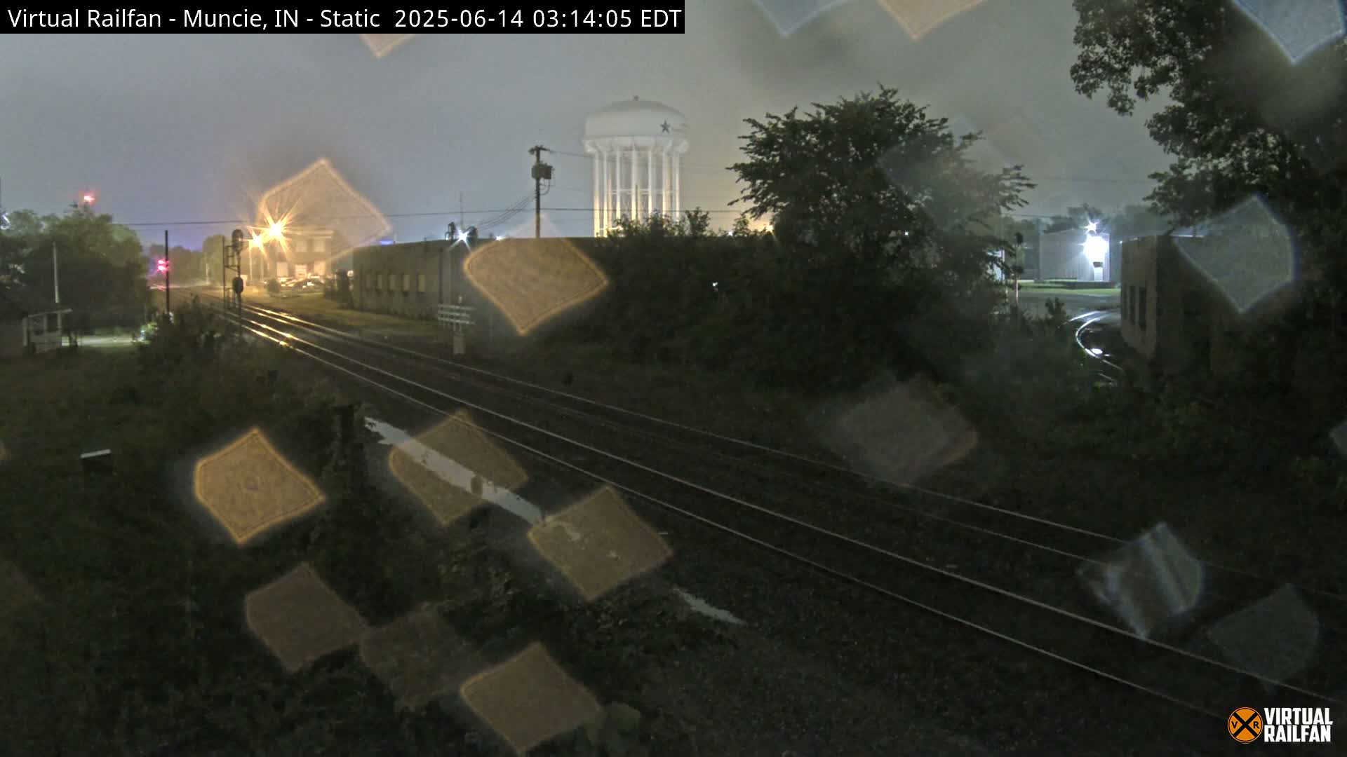 A nighttime view of railroad tracks, with rain obscuring the view, shows a water tower in the background.