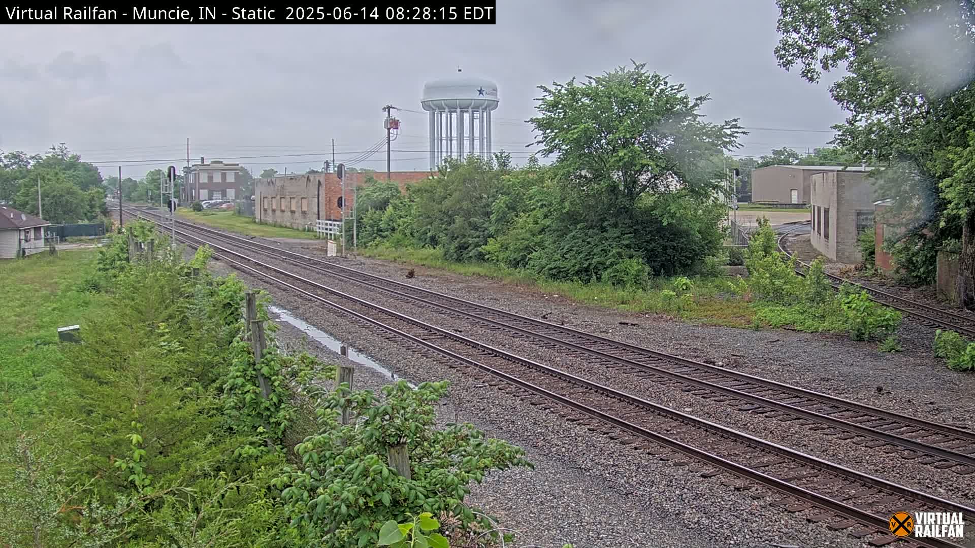 Overcast skies loom over a railway track that stretches into the distance, passing by industrial buildings and lush greenery, with a water tower visible in the background.