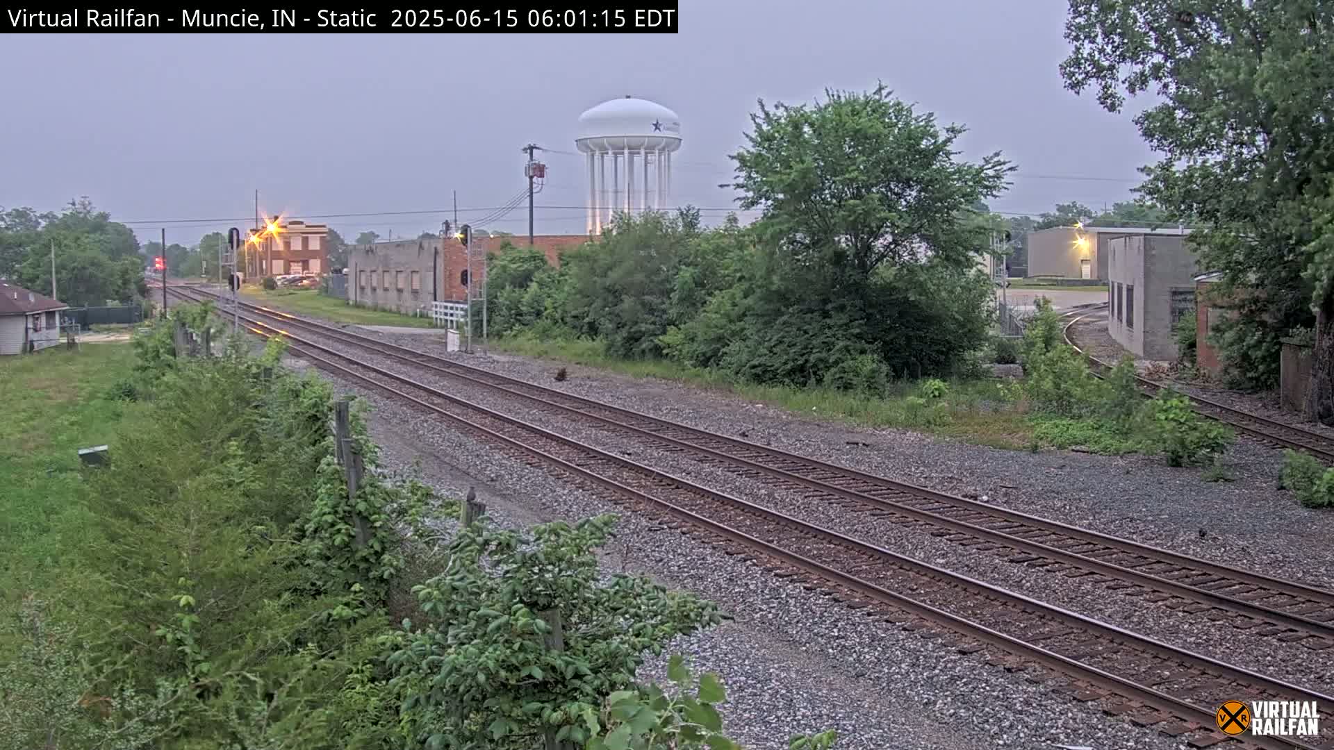 Triple railroad tracks stretch into the distance past industrial buildings and a water tower under an overcast sky.