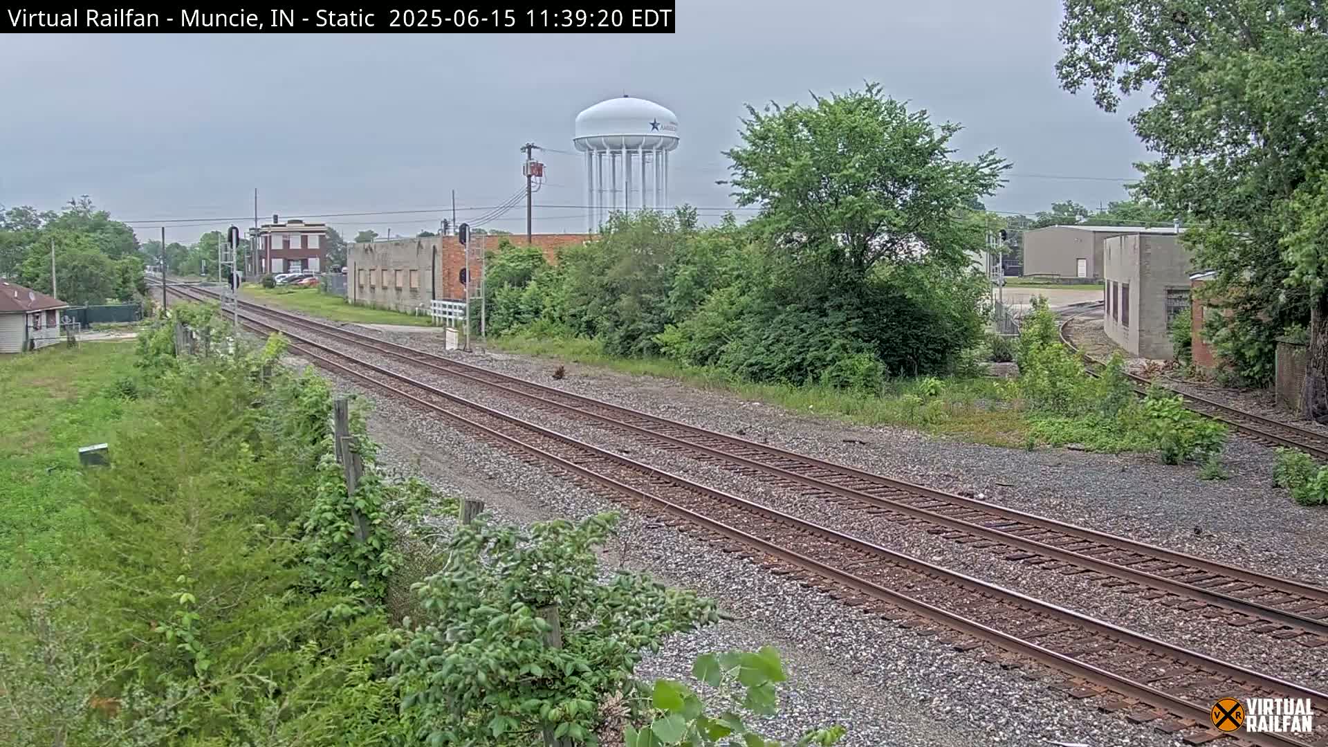 A view of railroad tracks extending into the distance under an overcast sky, passing industrial buildings and a water tower.