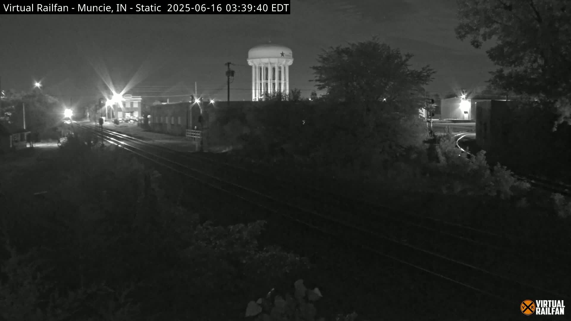 A nighttime, low-light view of railroad tracks leading toward a water tower and buildings under a partly cloudy sky.