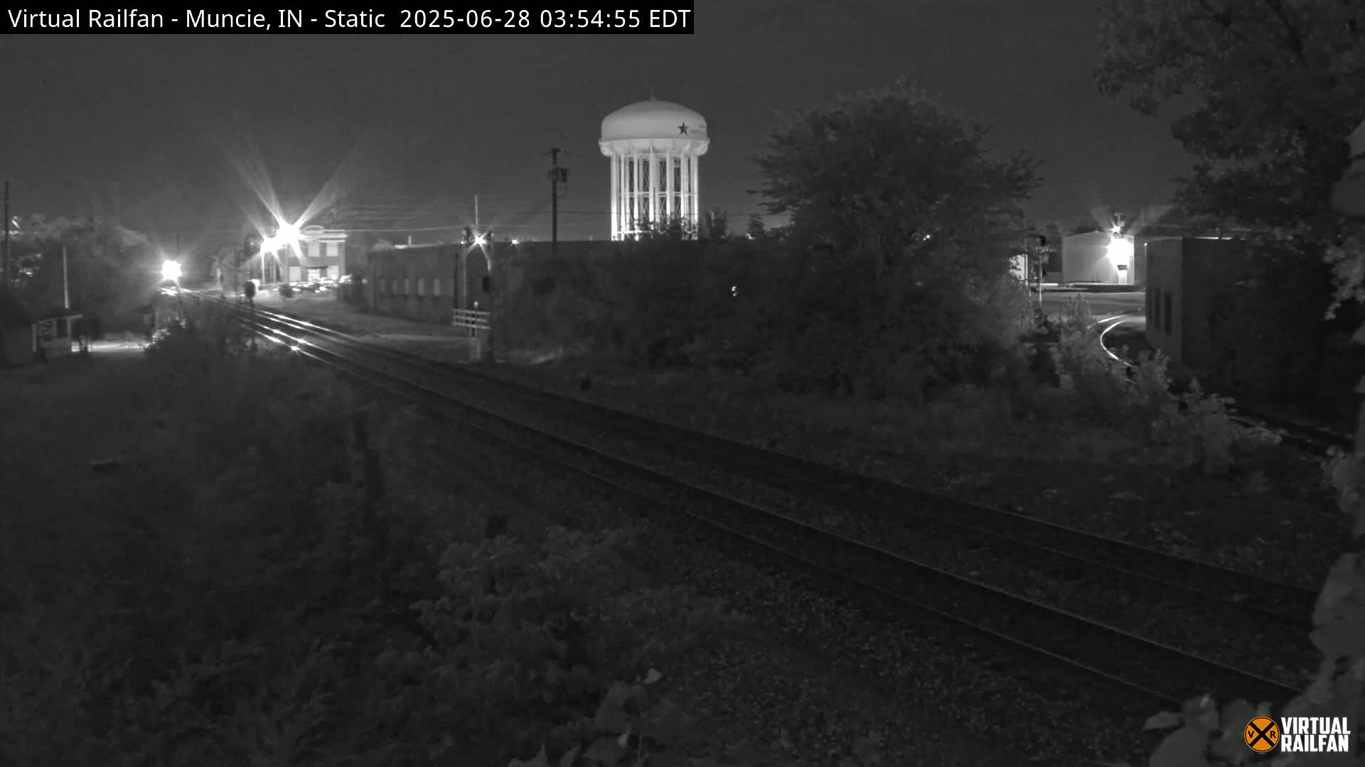 A nighttime view of railroad tracks leading toward a water tower and buildings under a dark sky.