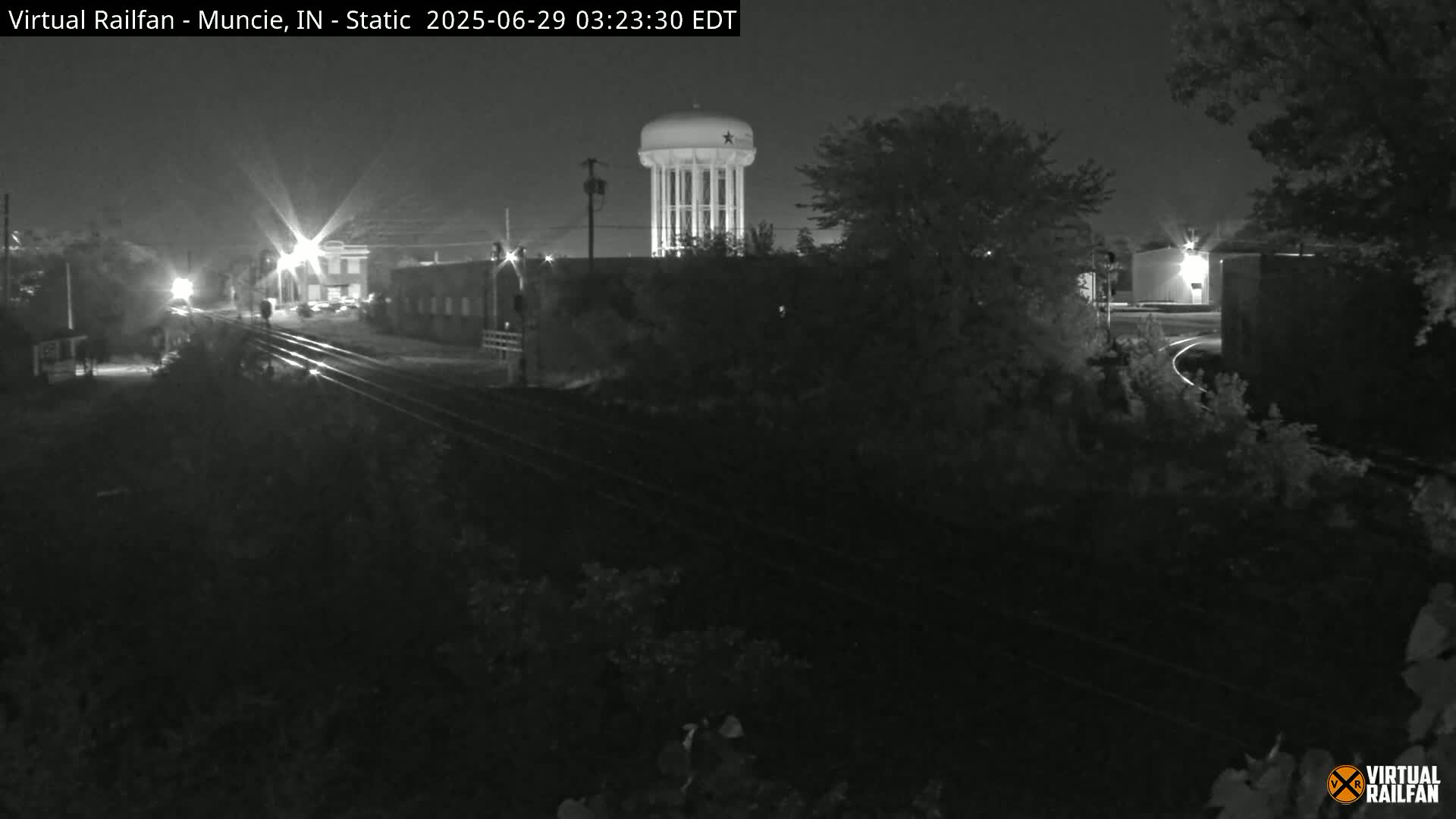 A nighttime, black-and-white view of railroad tracks and a water tower, with surrounding buildings and vegetation partially obscured by darkness.