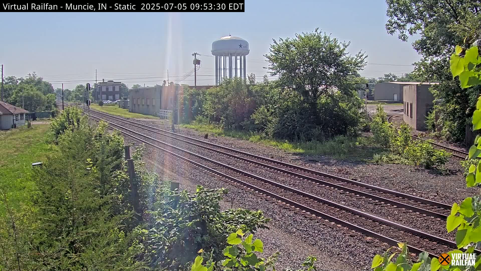 Three sets of train tracks extend into the distance under a sunny sky, passing alongside industrial buildings and a water tower.