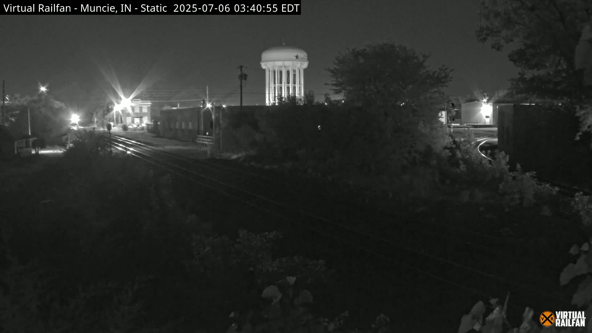 A nighttime view of railroad tracks leading toward a water tower and buildings under a dark sky.