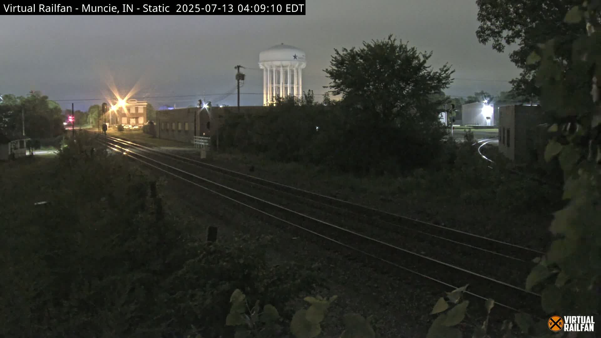 A nighttime view of a railroad track leading towards a water tower and some buildings under a dark, overcast sky.