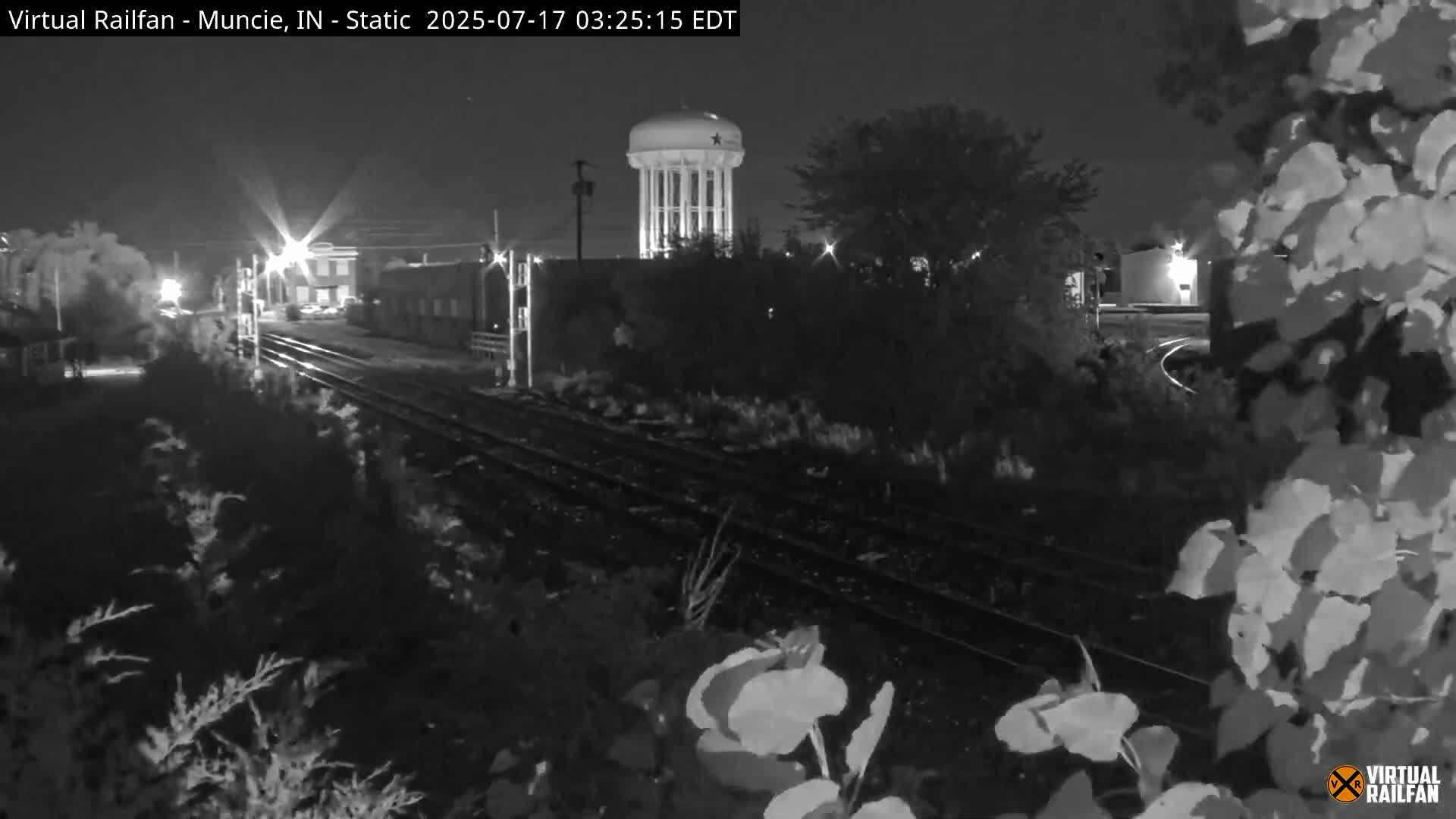 A nighttime view of railroad tracks with a water tower in the background, partially obscured by vegetation.