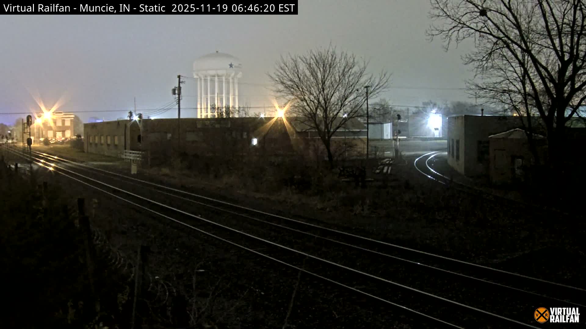 A nighttime view of railroad tracks with a water tower in the background, partially obscured by vegetation.