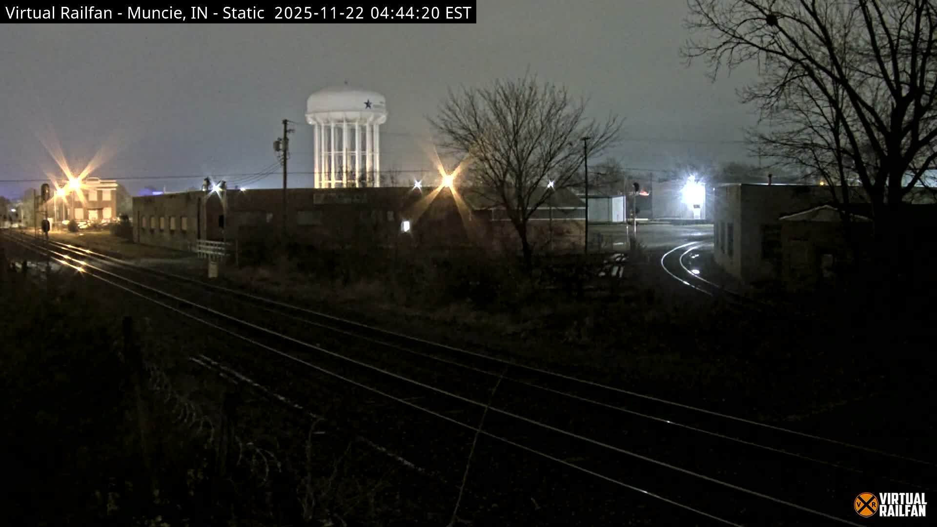 A nighttime scene captures multiple train tracks curving through an industrial area with bare trees and a brightly lit water tower under an overcast and damp sky, illuminated by various artificial lights.