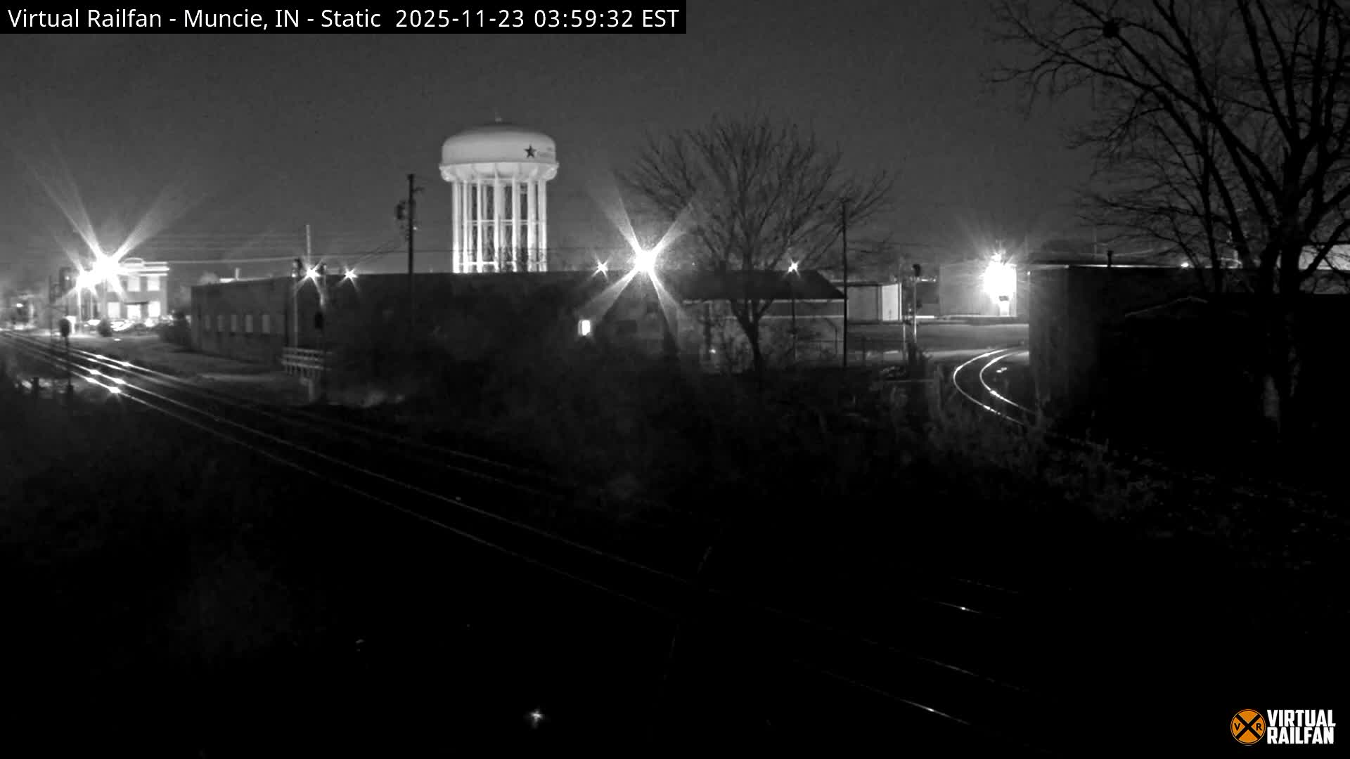 A nighttime view reveals multiple train tracks stretching into the distance past illuminated buildings, a prominent water tower, and bare trees under a clear, dark sky.
