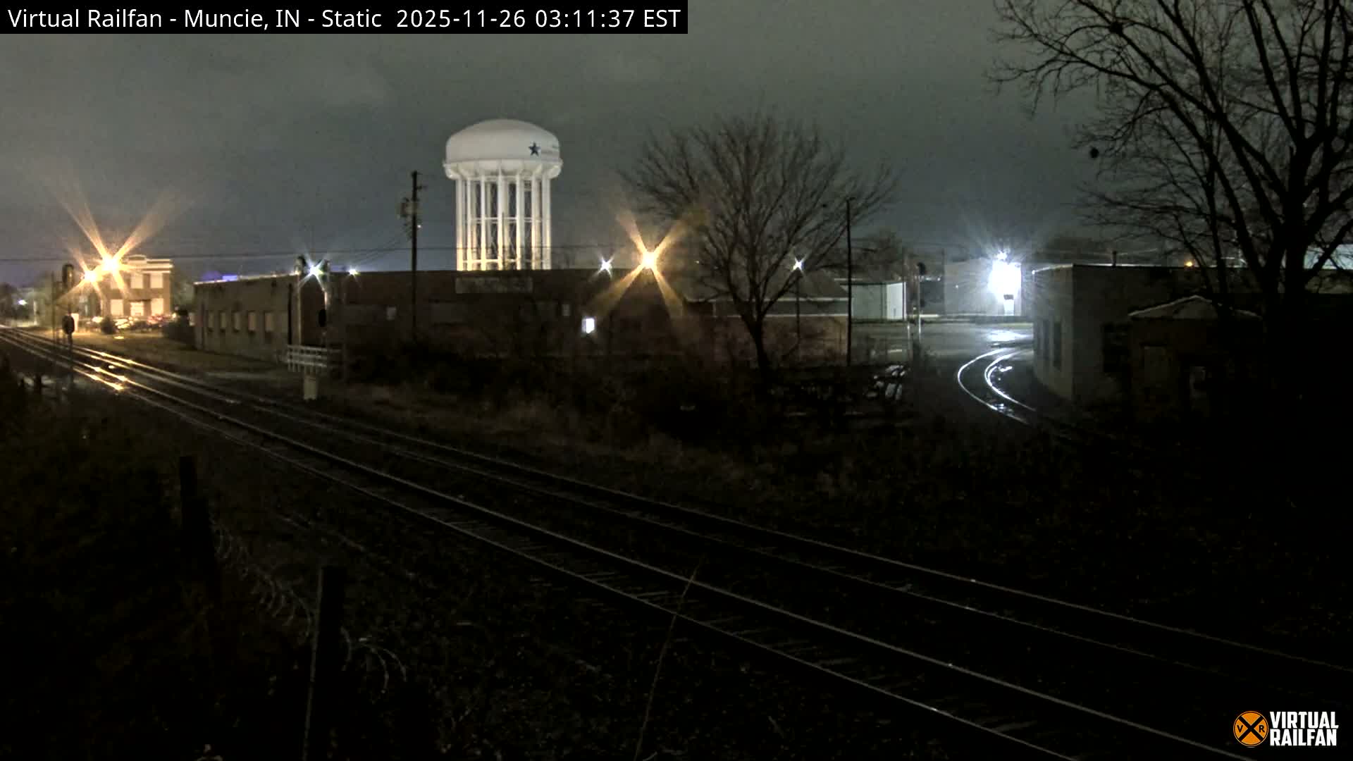 A nighttime view shows railroad tracks winding through a damp industrial area featuring buildings, bare trees, and a prominent water tower under an overcast sky.