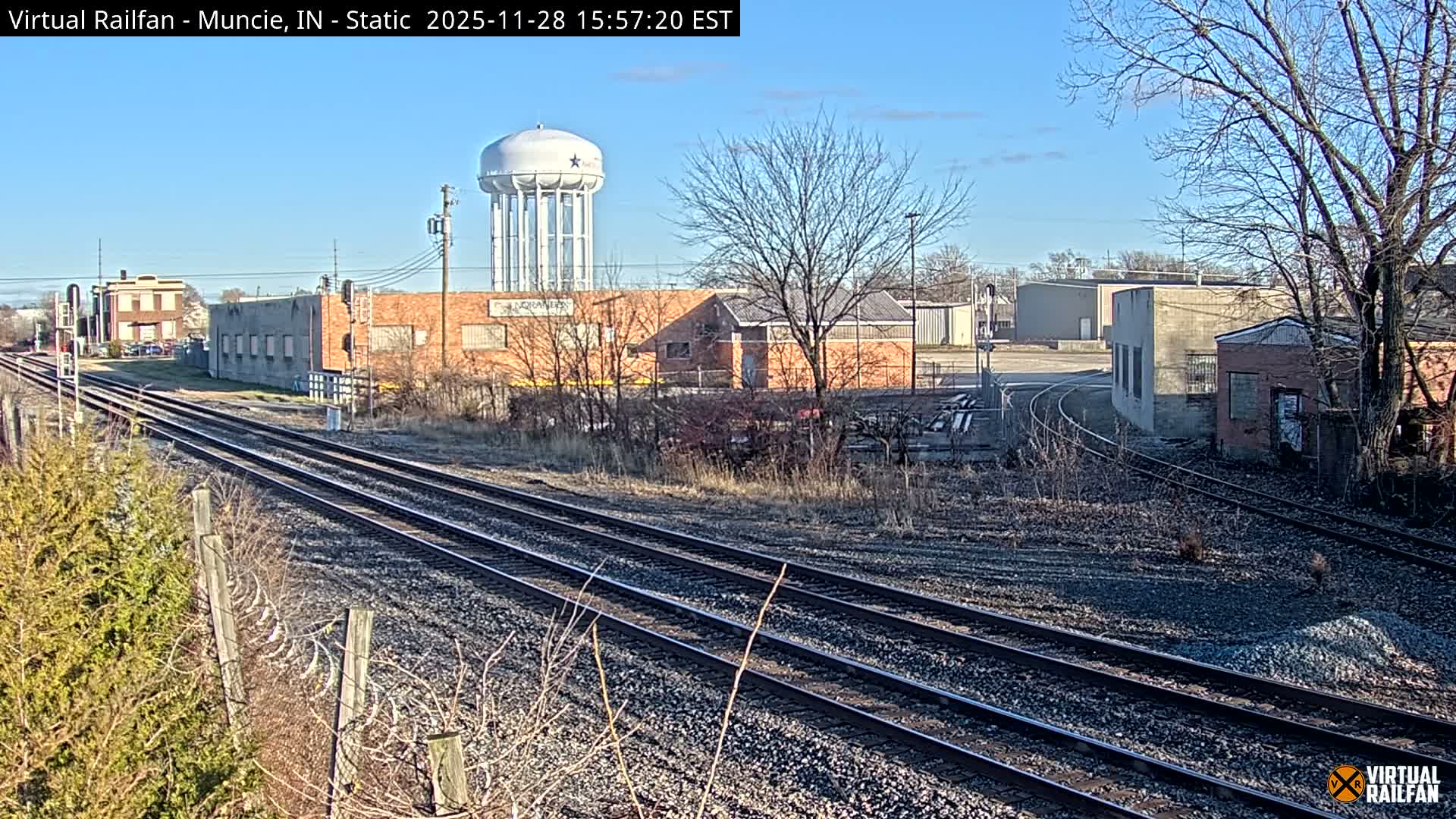 Multiple railroad tracks extend through an industrial landscape featuring various buildings, bare trees, and a distant water tower under a clear, sunny sky.
