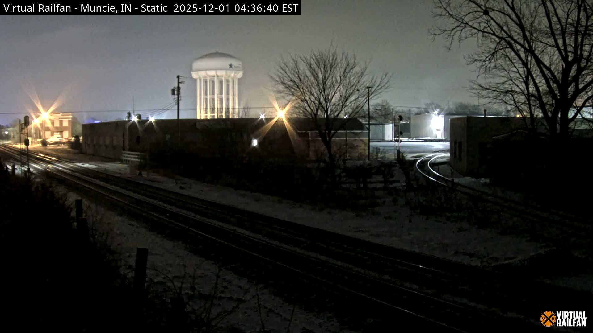 A light dusting of snow covers the ground around multiple railroad tracks, bare trees, and illuminated industrial buildings, with a prominent water tower standing against the dark night sky.