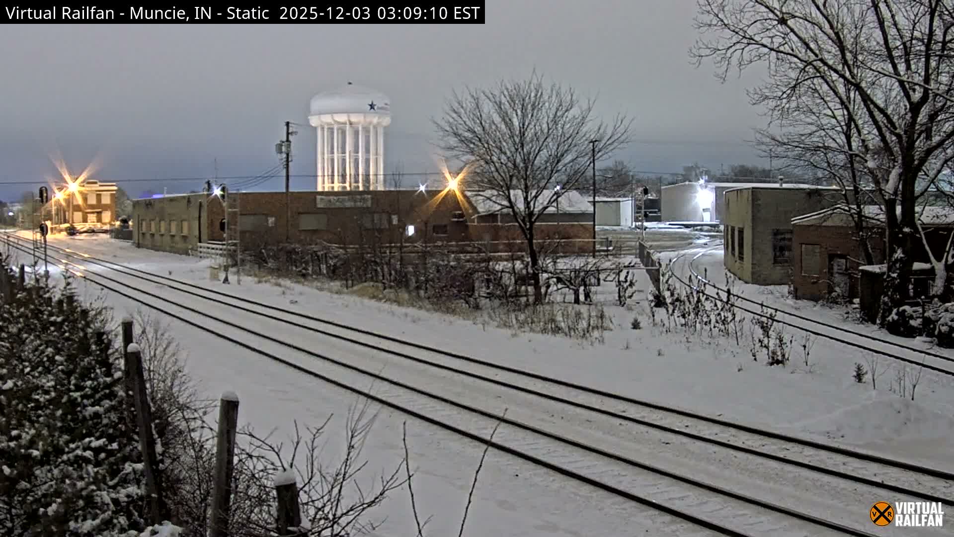 A snow-covered landscape under a dark, overcast sky features multiple railroad tracks leading through an industrial area with various buildings and bare trees, all illuminated by scattered artificial lights, with a tall water tower standing in the distance.