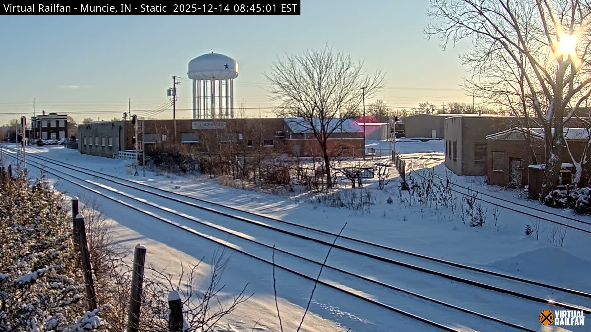 A snow-covered landscape under a dark, overcast sky features multiple railroad tracks leading through an industrial area with various buildings and bare trees, all illuminated by scattered artificial lights, with a tall water tower standing in the distance.