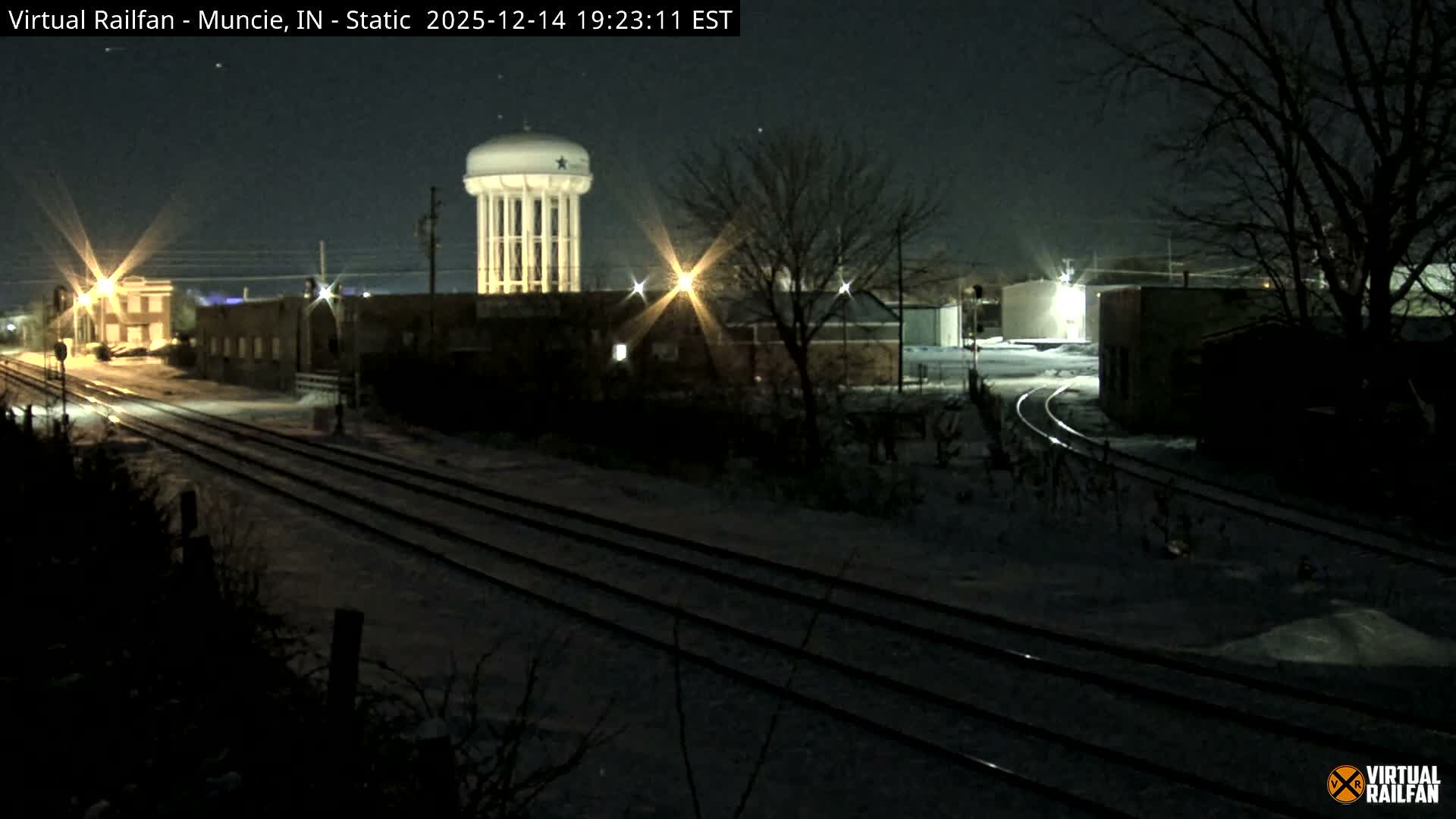 A snow-covered landscape under a dark, overcast sky features multiple railroad tracks leading through an industrial area with various buildings and bare trees, all illuminated by scattered artificial lights, with a tall water tower standing in the distance.