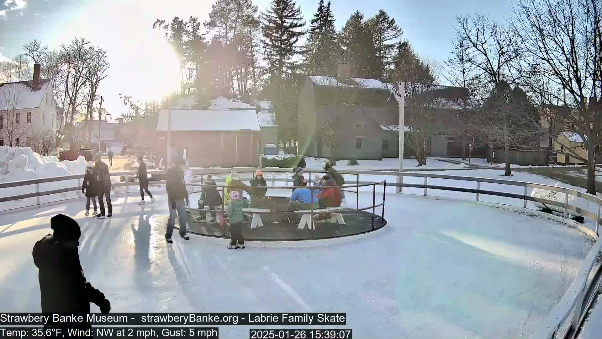 A group of people ice skate on a rink outdoors on a sunny, but cold, day with a light northwest wind.