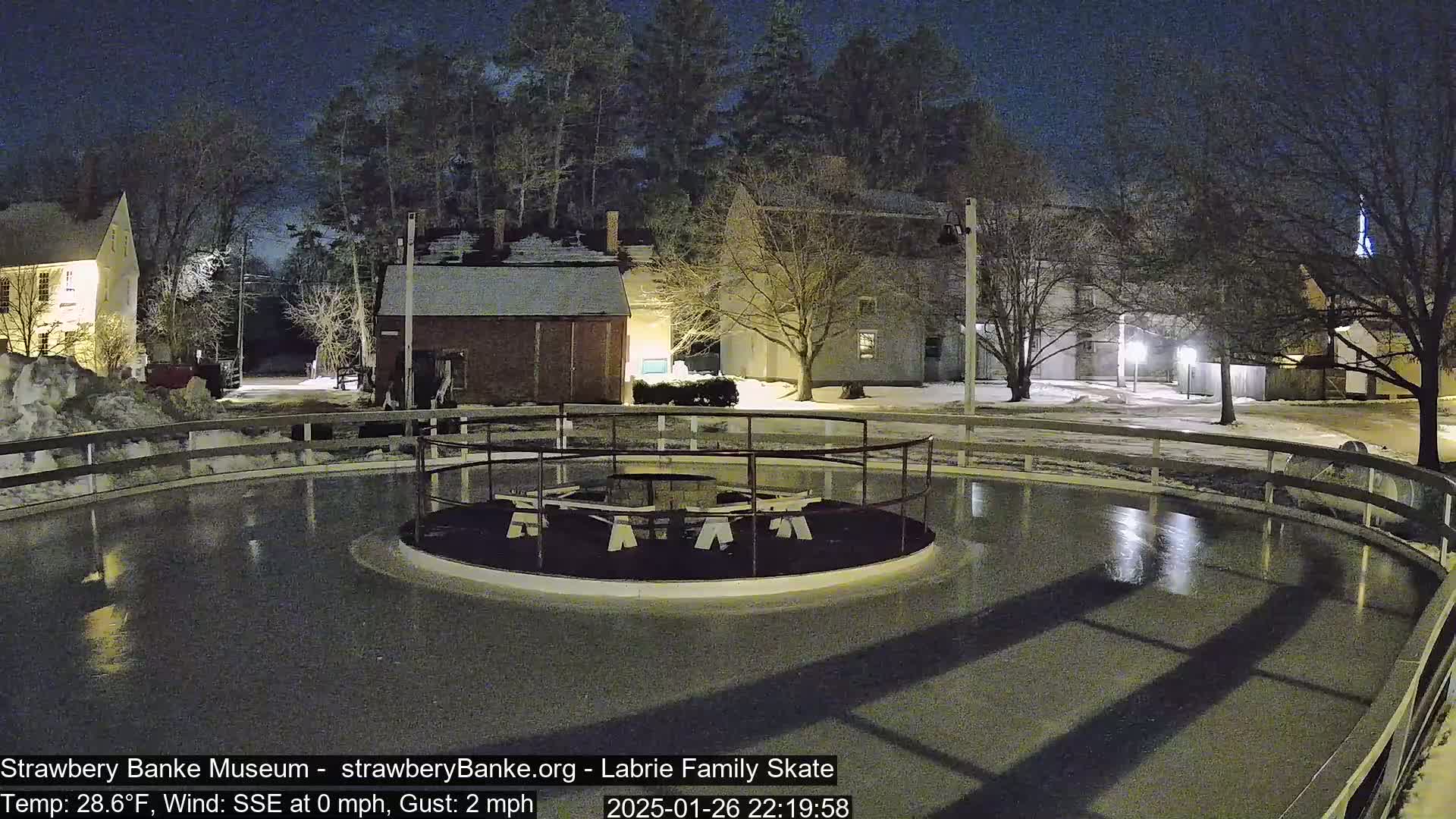 An outdoor ice skating rink at night, illuminated by lights, is surrounded by snow-covered ground and buildings under a dark sky with a slight wind.
