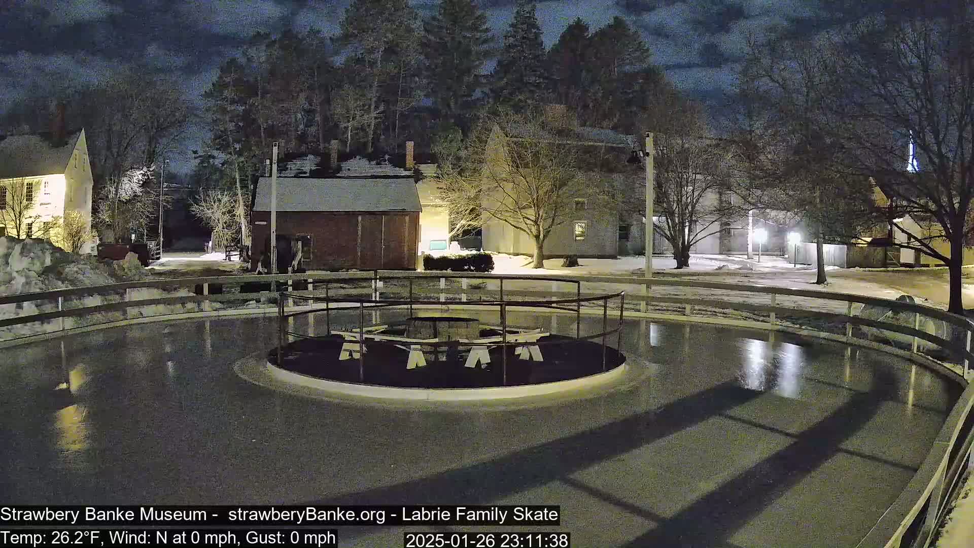 A nighttime view of an outdoor ice skating rink, surrounded by snow-covered ground and buildings under a partly cloudy sky.