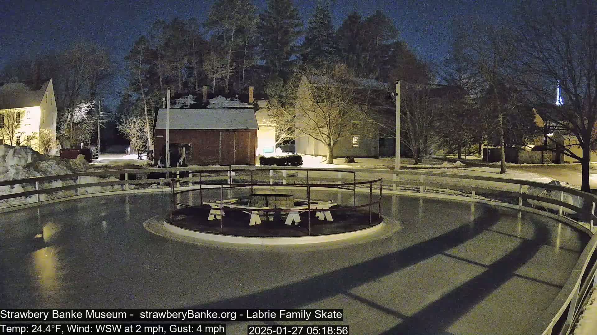 Here is a description of the image: An outdoor, nighttime view of an ice skating rink surrounded by a wooden fence, with several buildings and bare trees in the background under a dark sky.