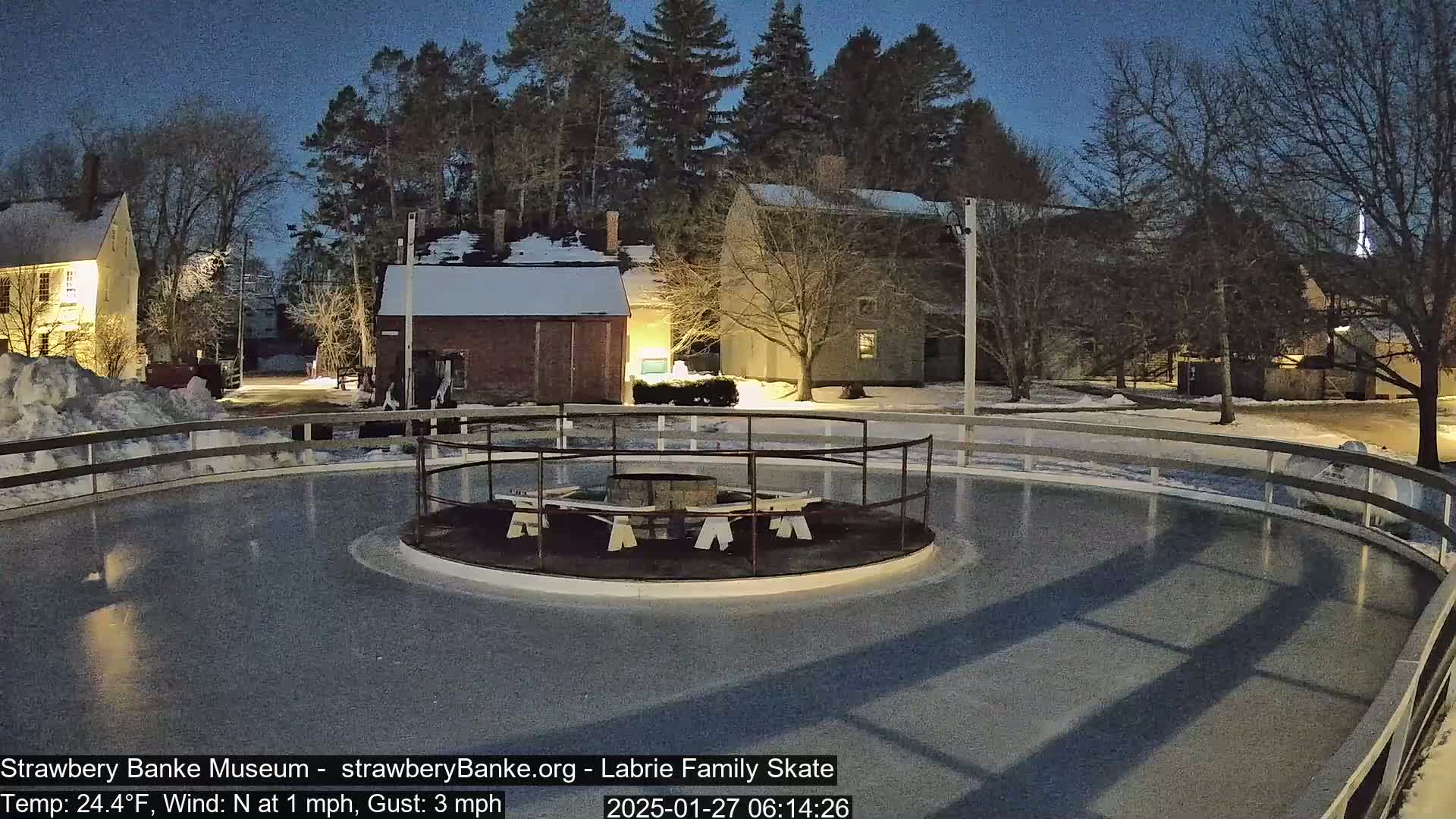 An outdoor ice skating rink is illuminated at night, surrounded by snow-covered buildings and trees under a twilight sky.
