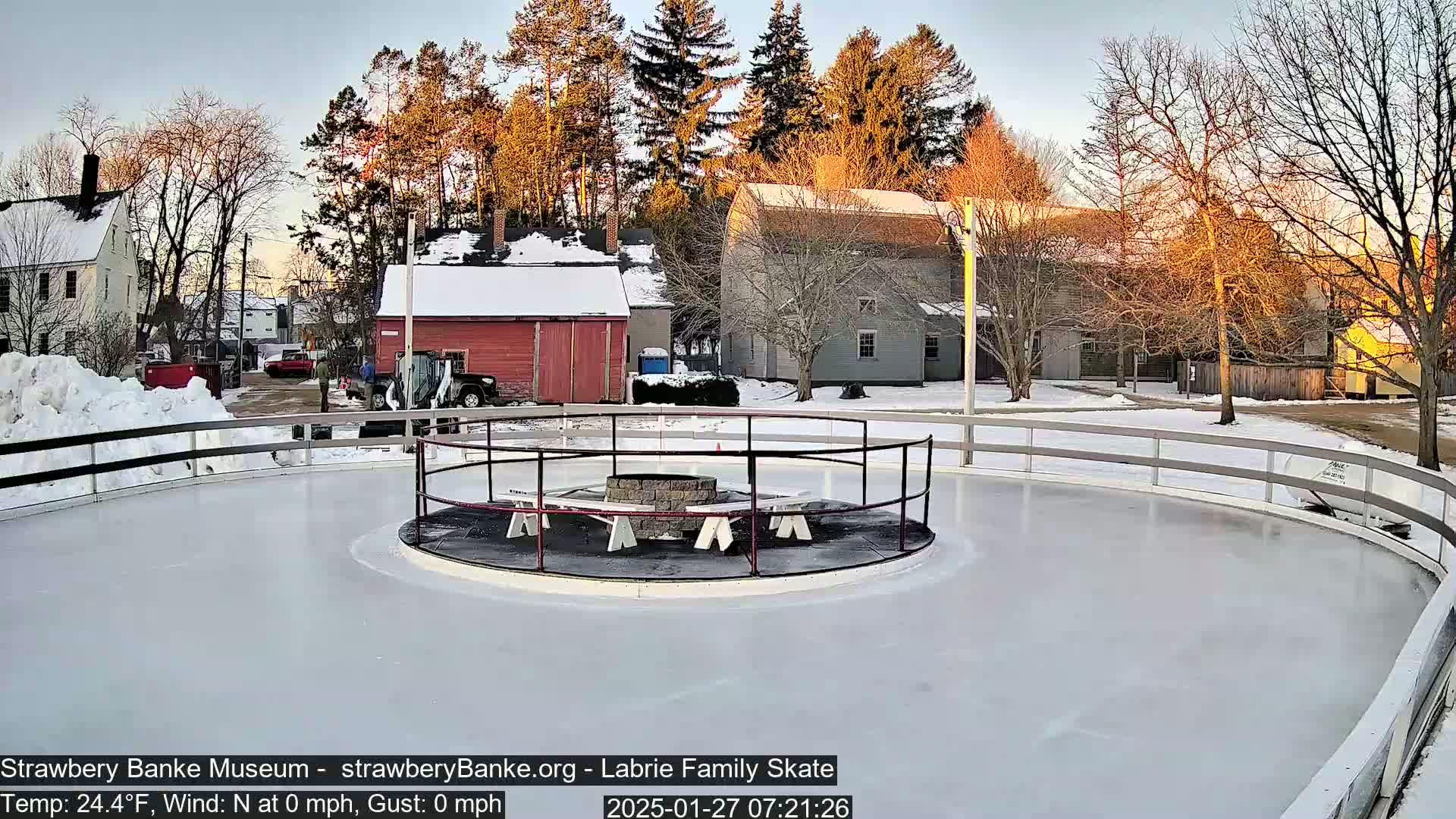 An outdoor ice skating rink with a fire pit in the center is surrounded by snow-covered buildings and trees under a twilight sky.