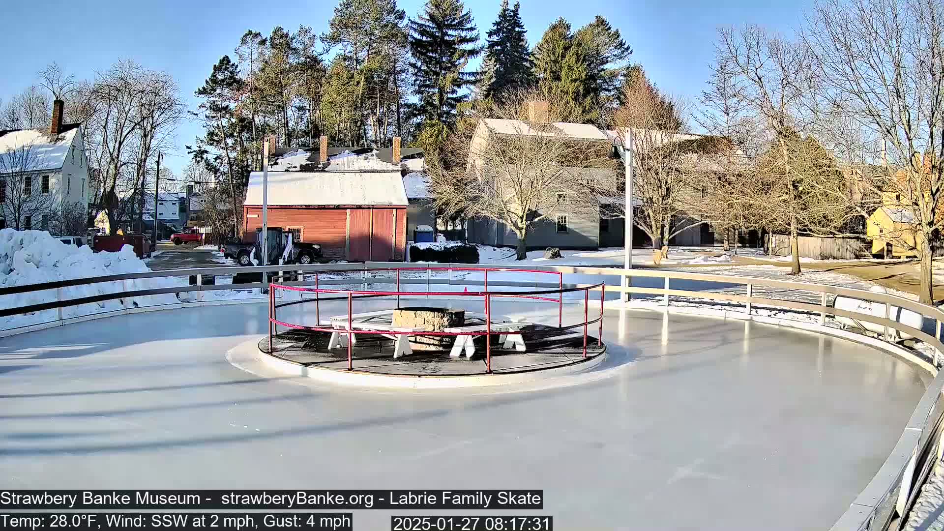 A circular ice rink, surrounded by a low wooden fence, is situated near several snow-covered buildings on a sunny winter day.