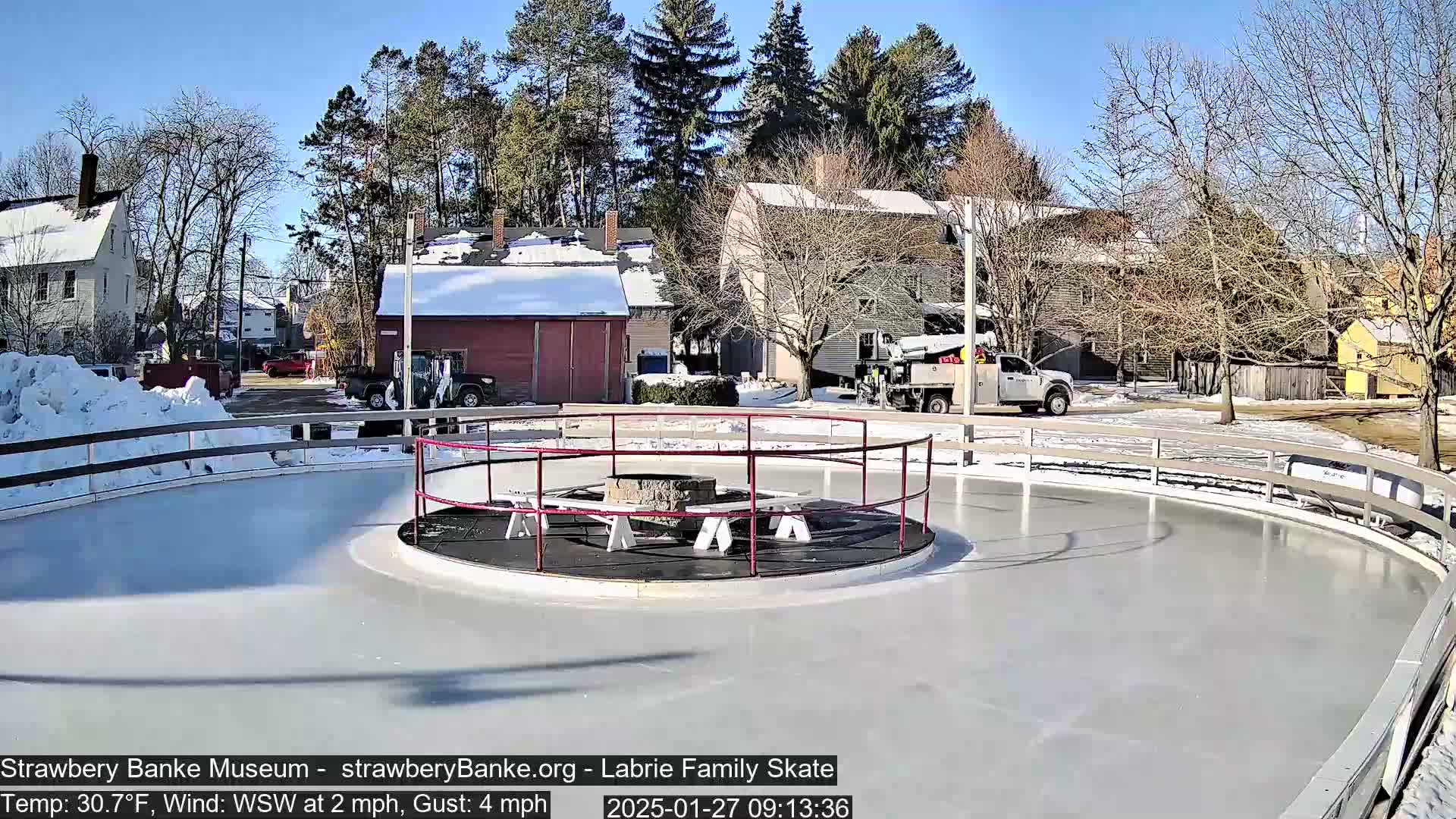 An outdoor ice skating rink, surrounded by a low fence and a fire pit in the center, is situated near several buildings under a sunny sky.