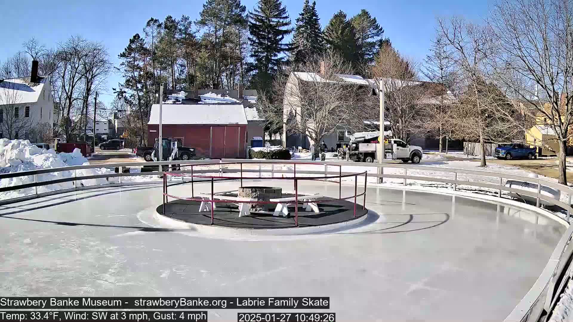 A sunny outdoor ice rink, surrounded by snow and buildings, features a central fire pit with seating.