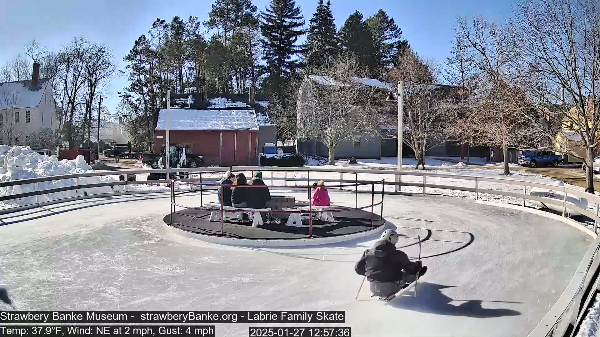 A person sleds on an outdoor ice rink surrounded by a low fence, with several people sitting on a bench in the center, under a sunny sky.