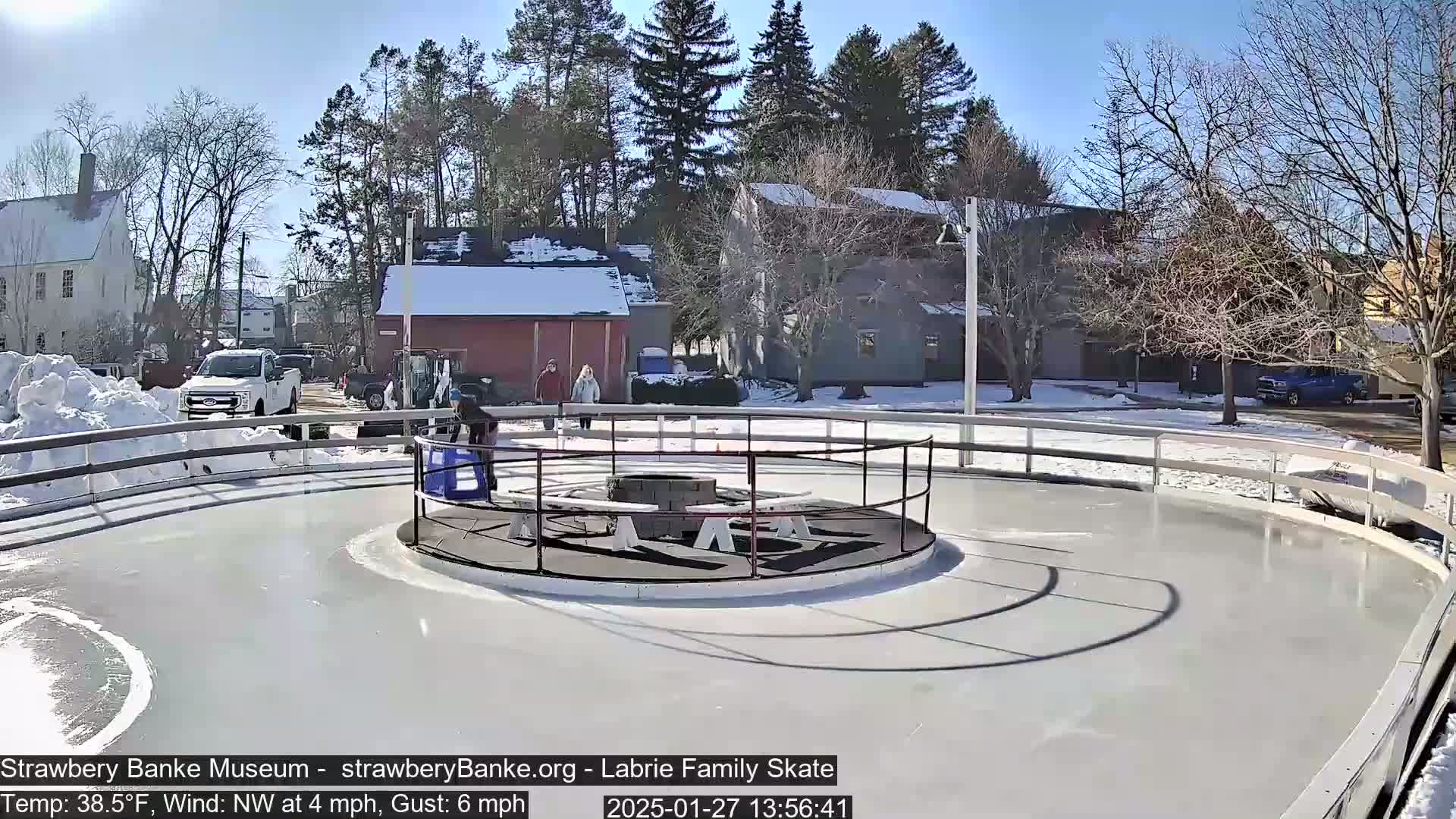 A sunny outdoor ice skating rink, surrounded by snow-covered ground and buildings, with several people near the center.