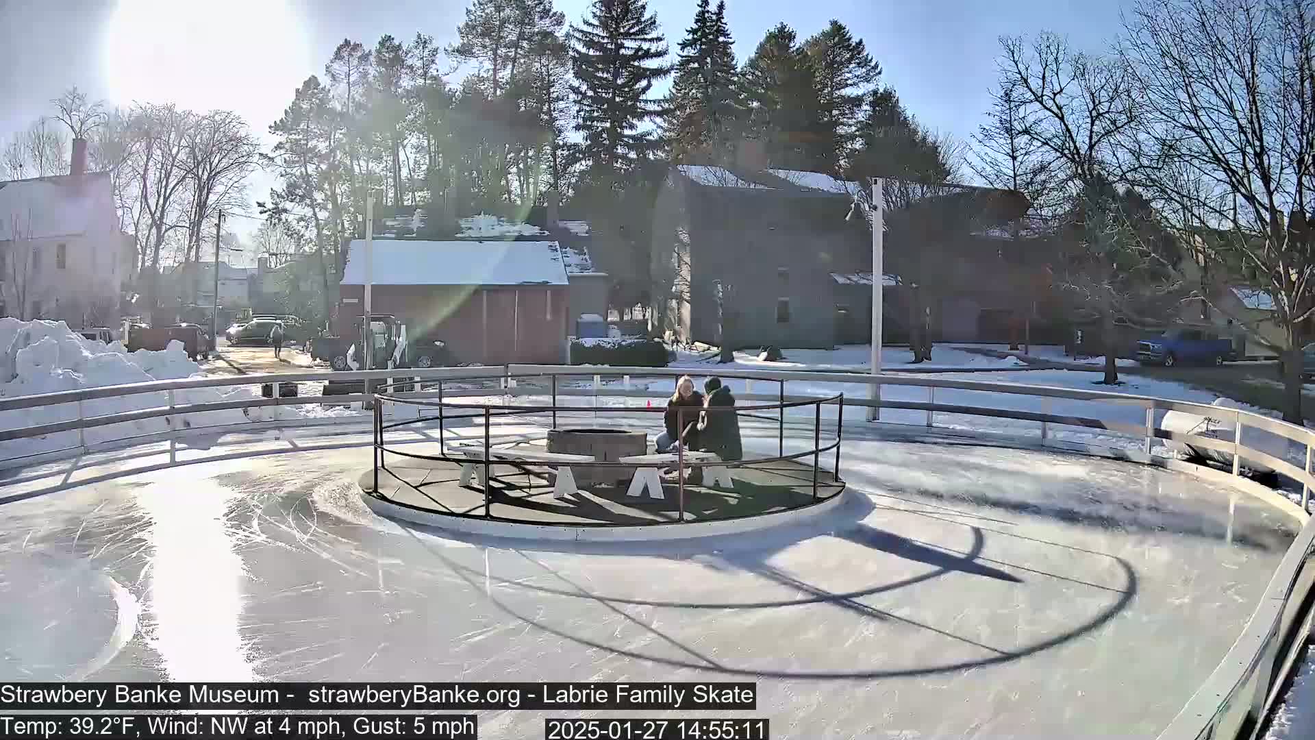A sunny day with a northwest wind shows two people sitting near a fire pit in the center of an outdoor ice skating rink, surrounded by snow-covered buildings and trees.