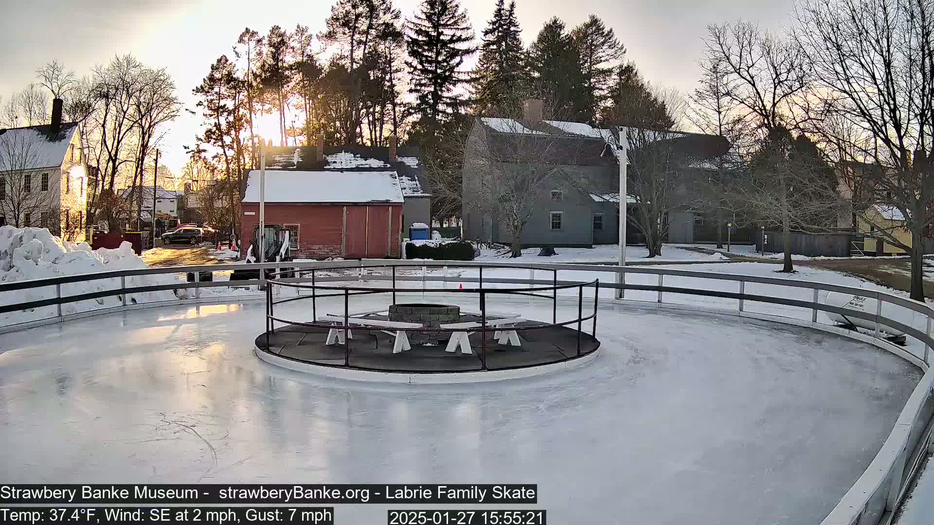 A snow-covered outdoor ice rink with a fire pit in the center is surrounded by buildings and trees under a partly sunny sky.