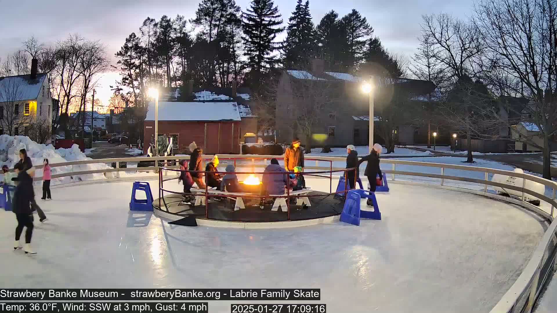 An outdoor ice skating rink at dusk, with people ice skating and gathered around a fire pit in the center, under a mostly clear sky.