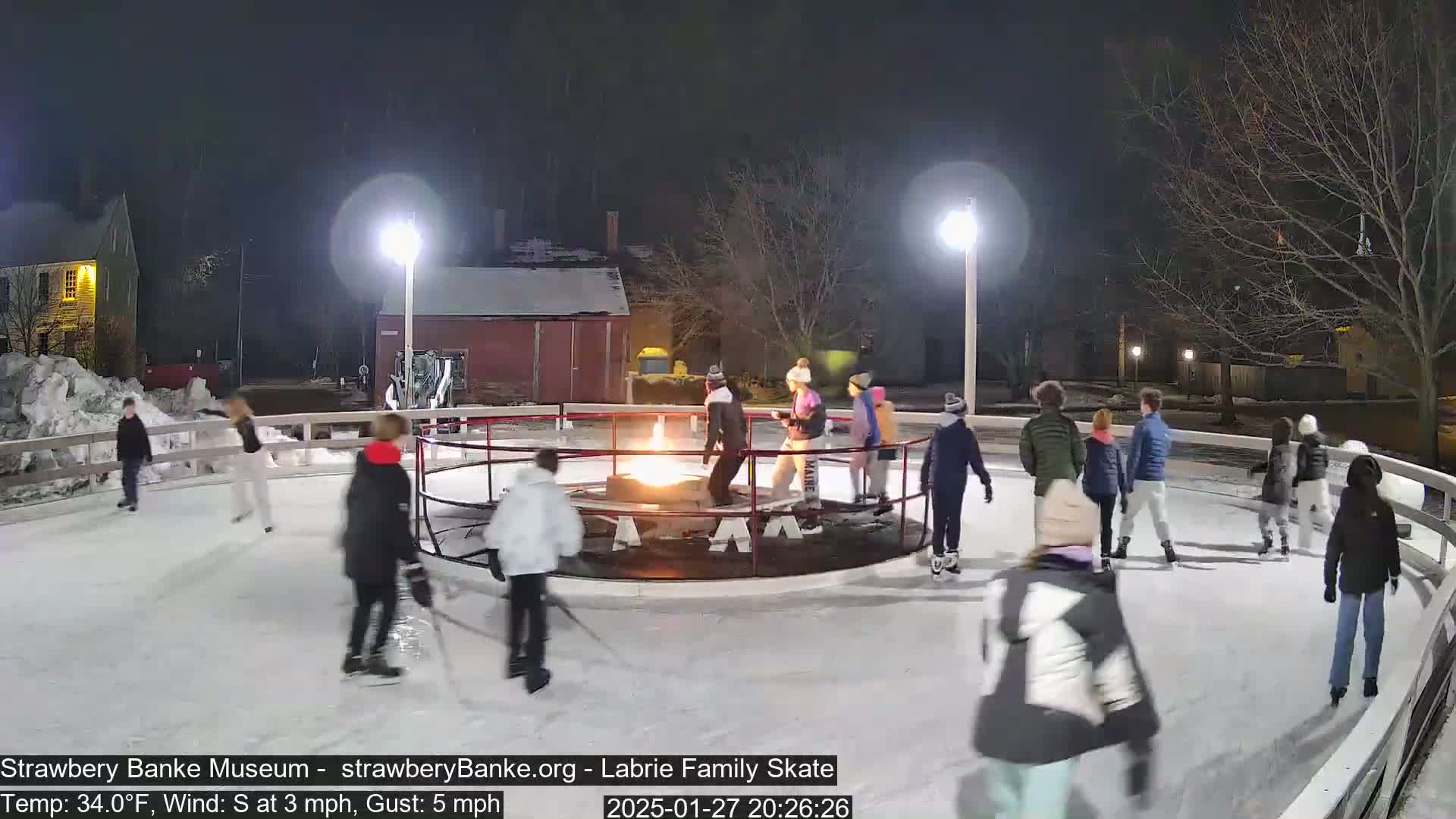 At night, under a dark sky, many people ice skate on an outdoor rink surrounding a fire pit.