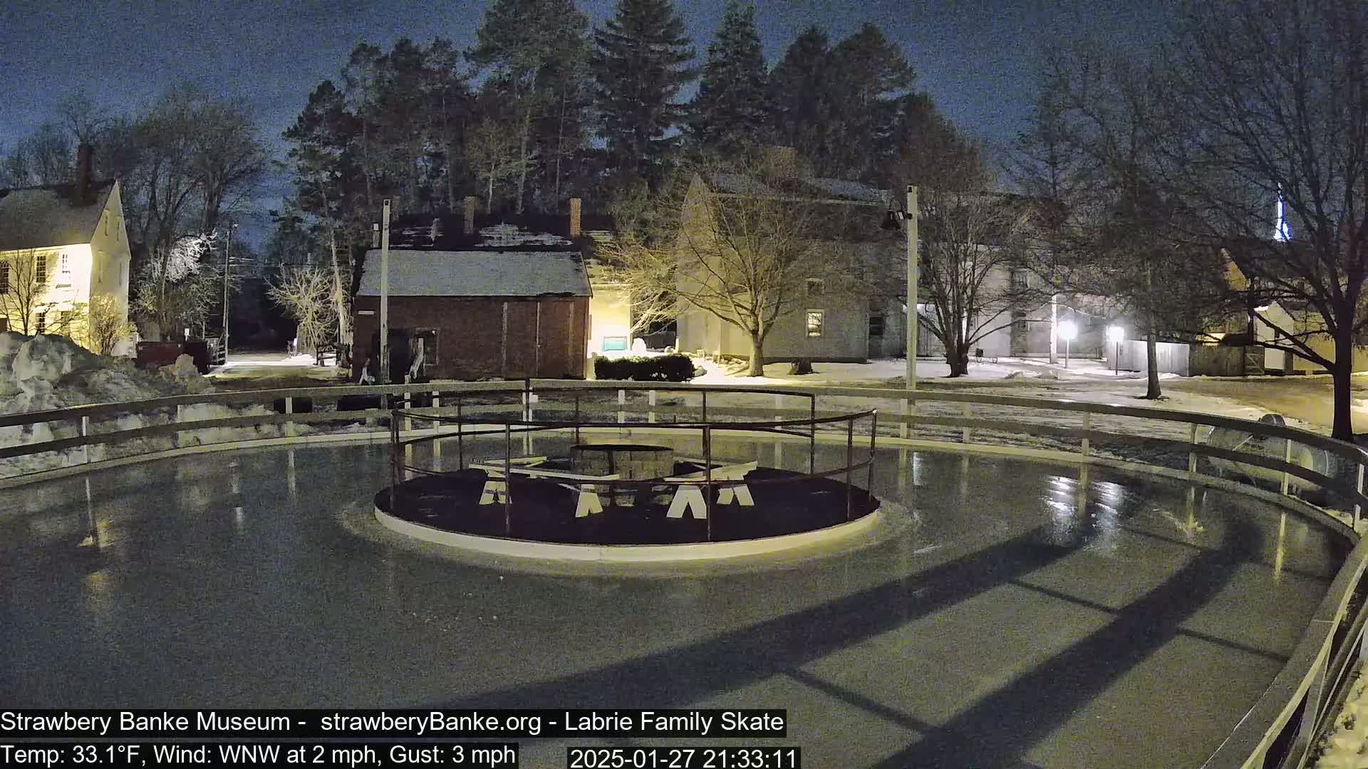 A nighttime view of an outdoor ice skating rink, illuminated, surrounded by snow-covered ground and buildings under a dark sky with a light wind.