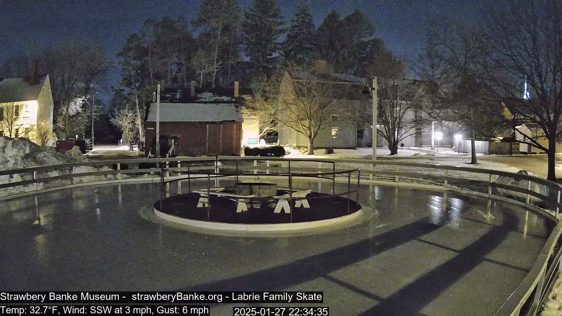 A nighttime view of an outdoor ice rink, surrounded by snow-covered ground and buildings under a dark sky.