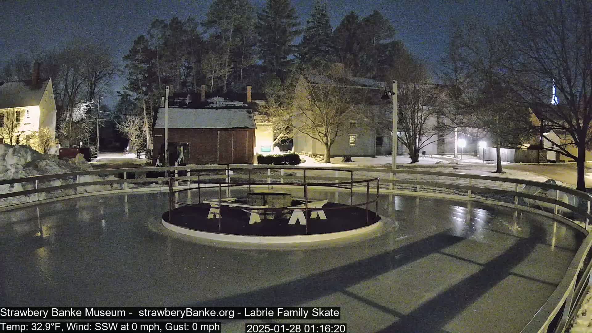 An outdoor ice skating rink, surrounded by snow-covered ground and illuminated buildings, is visible at night under a dark sky.