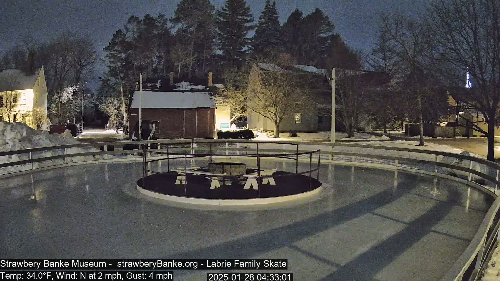 A nighttime view of a snow-covered outdoor ice skating rink with a fire pit in the center, surrounded by buildings and trees under a dark sky.