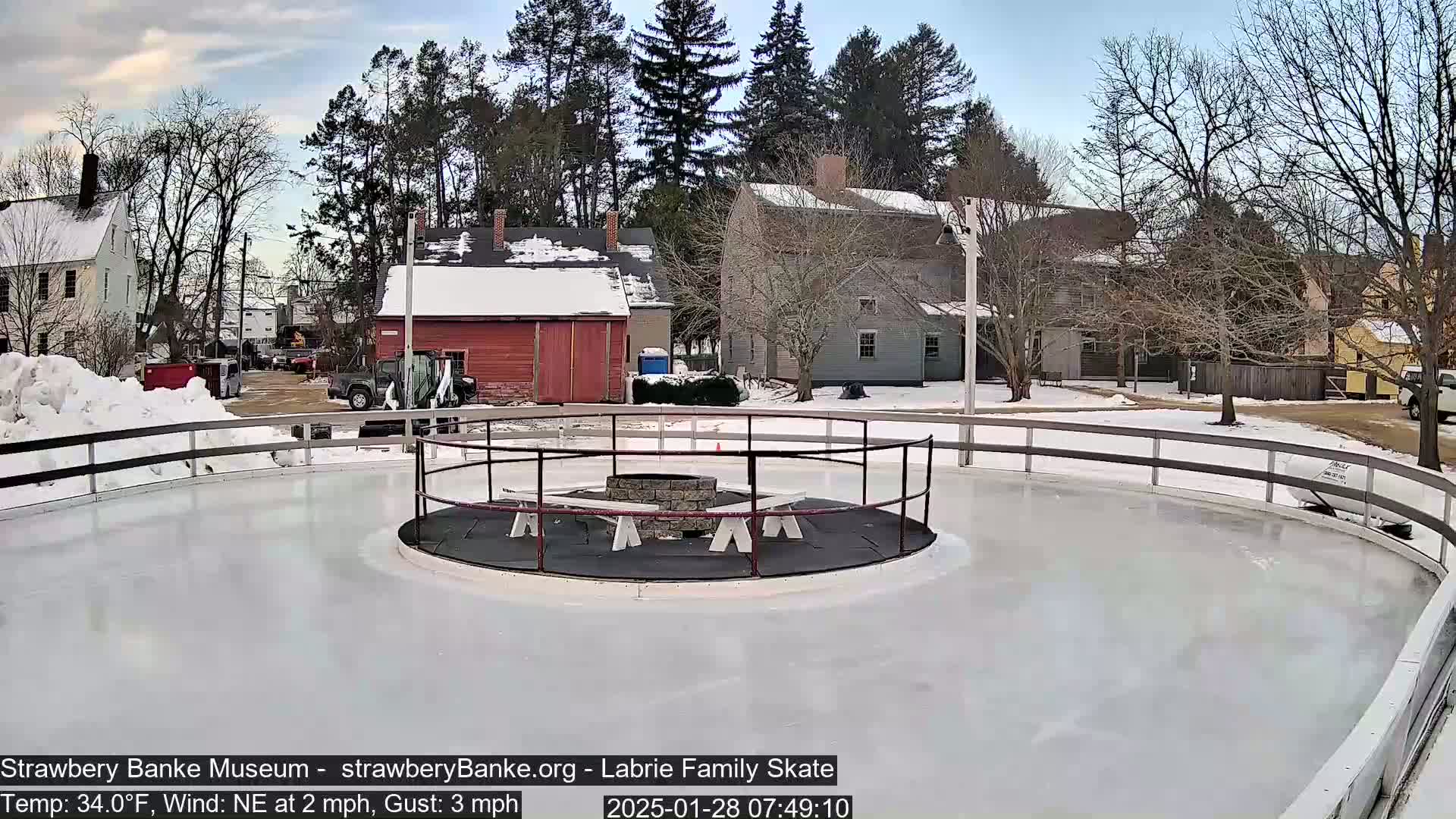 A circular ice skating rink with a fire pit in the center is surrounded by a snow-covered landscape and buildings, under a mostly clear sky.