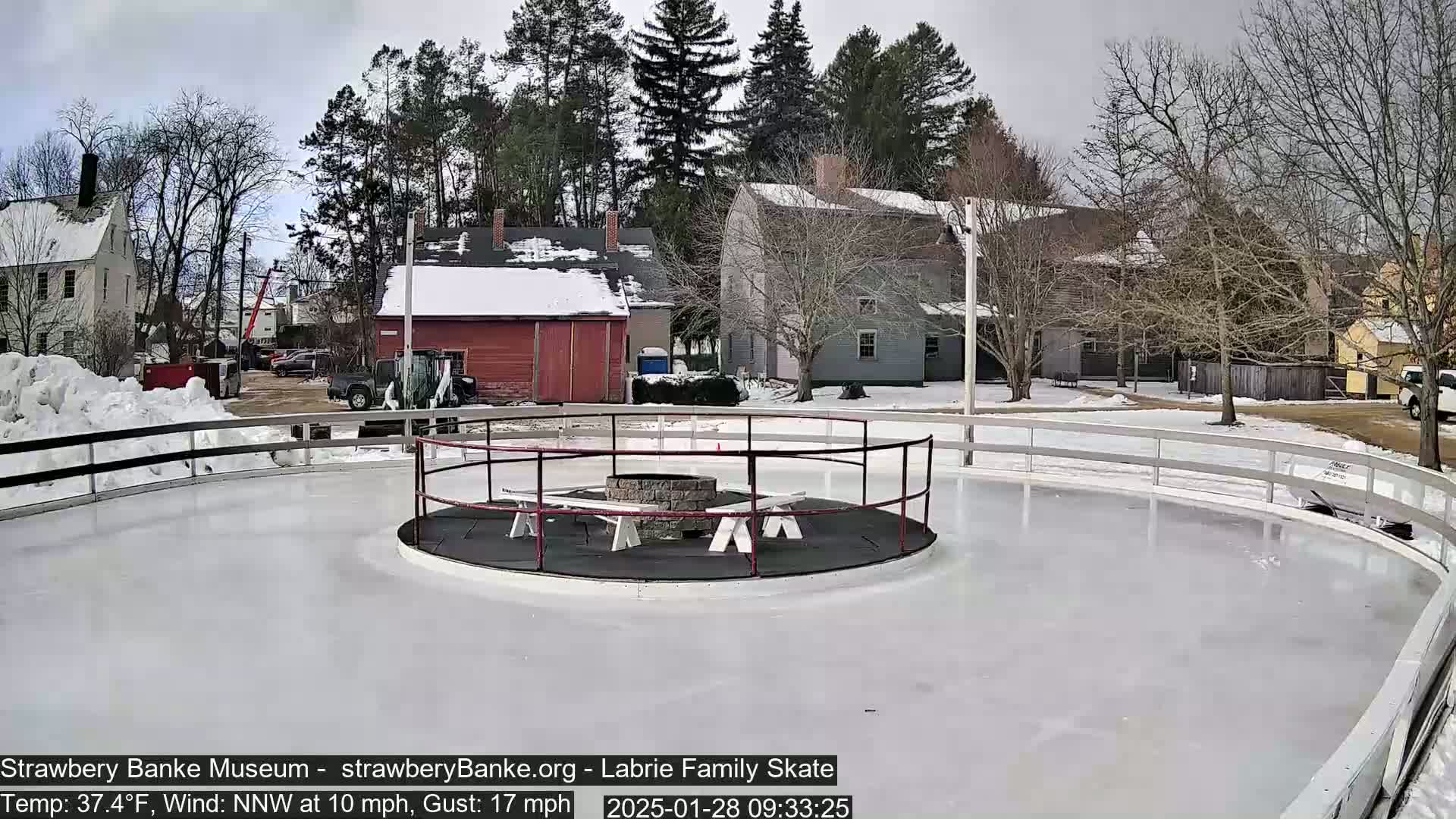 A circular ice skating rink with a fire pit and benches in the center is surrounded by a snow-covered landscape and several buildings under an overcast sky.
