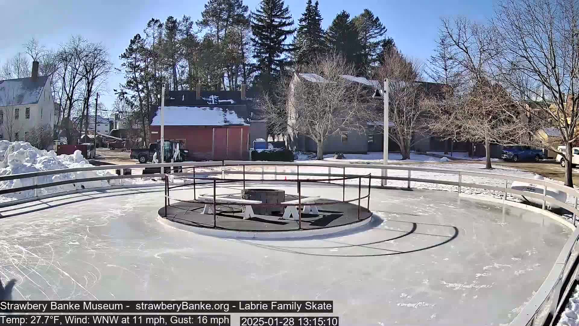 A sunny outdoor ice rink with a fire pit in the center, surrounded by buildings and snow-covered ground.